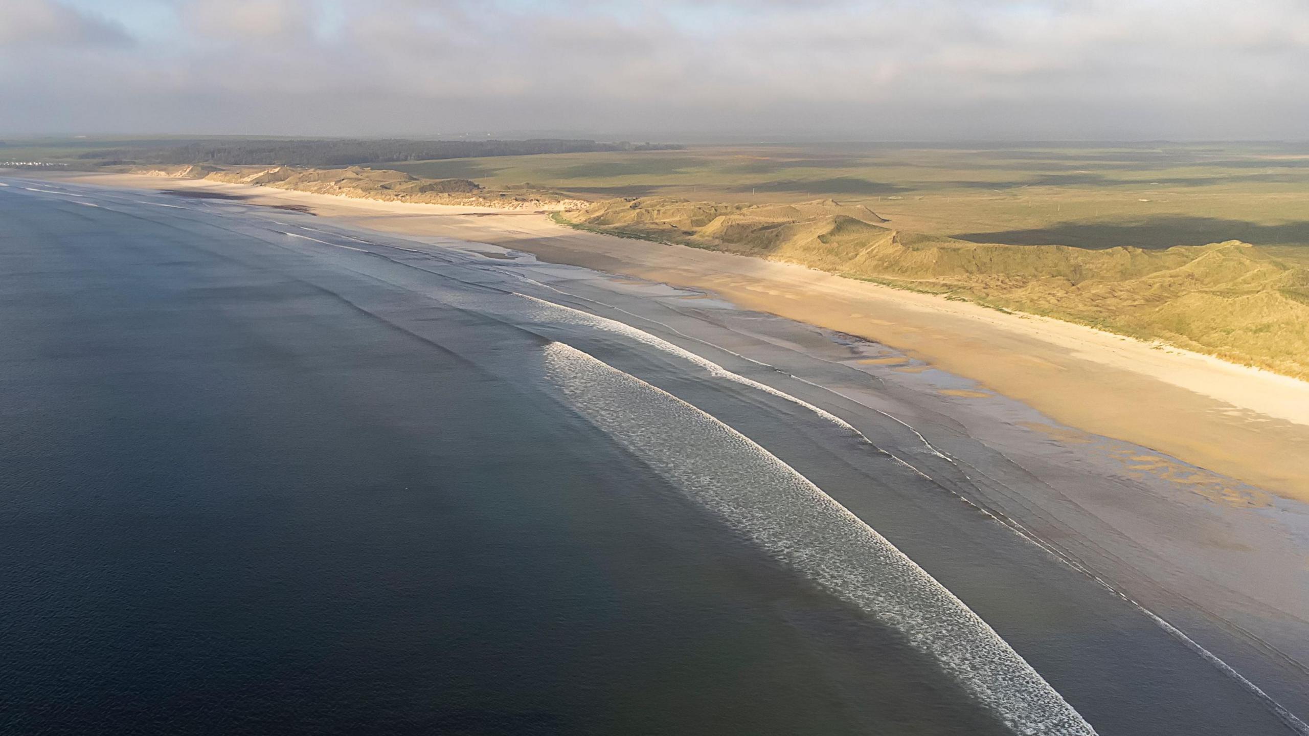 A stretch of Dunnet Bay with golden sands and waves lapping the shore viewed from above