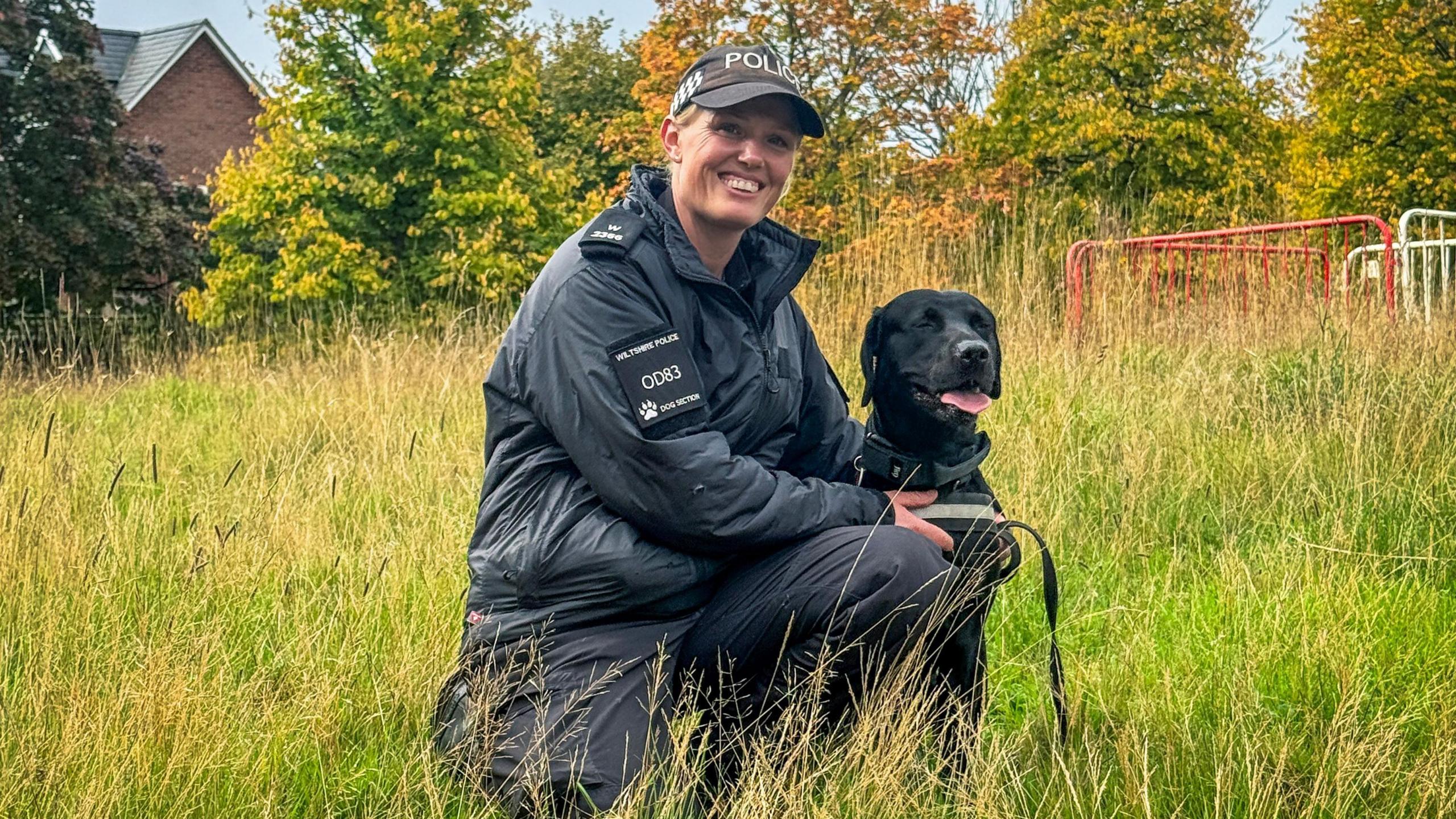 Woman in black police jacket and wearing a hat which reads 'POLICE'. She is holding a black dog with its tongue out and they are in grass.