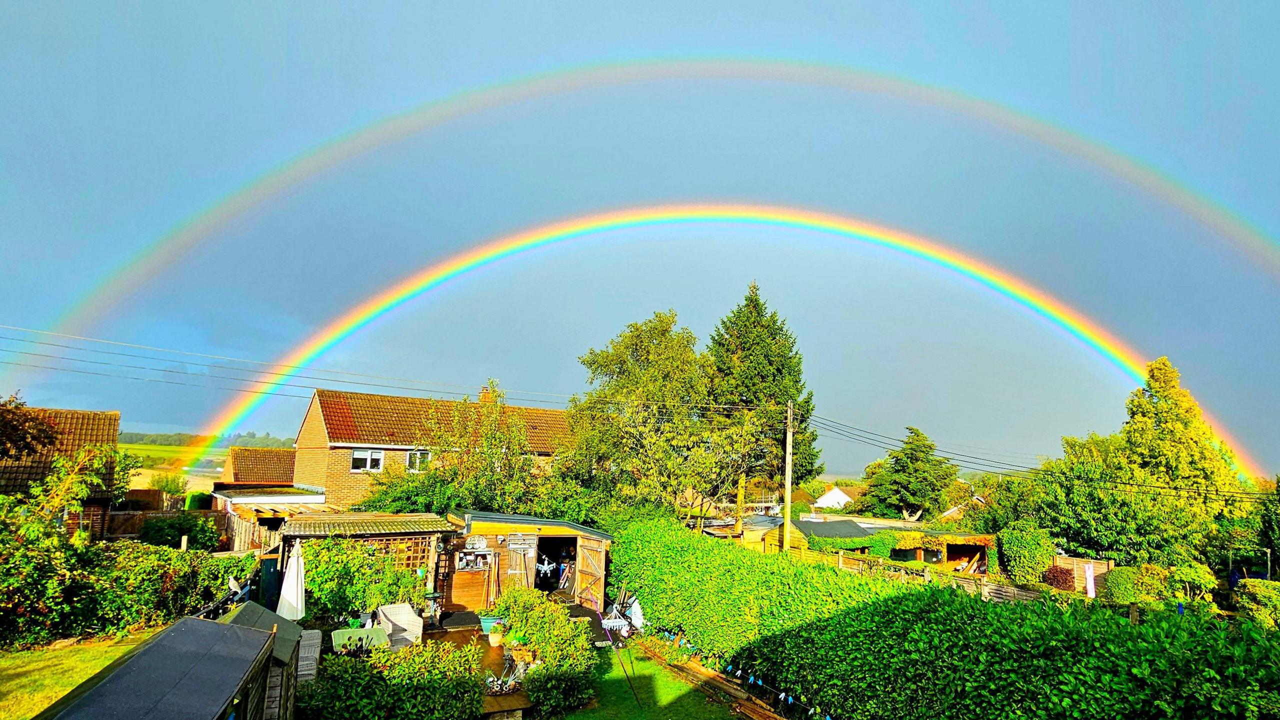 The sun is shining after heavy rain across a garden seen with neighbouring houses in the background. Above it all shine two full rainbows.