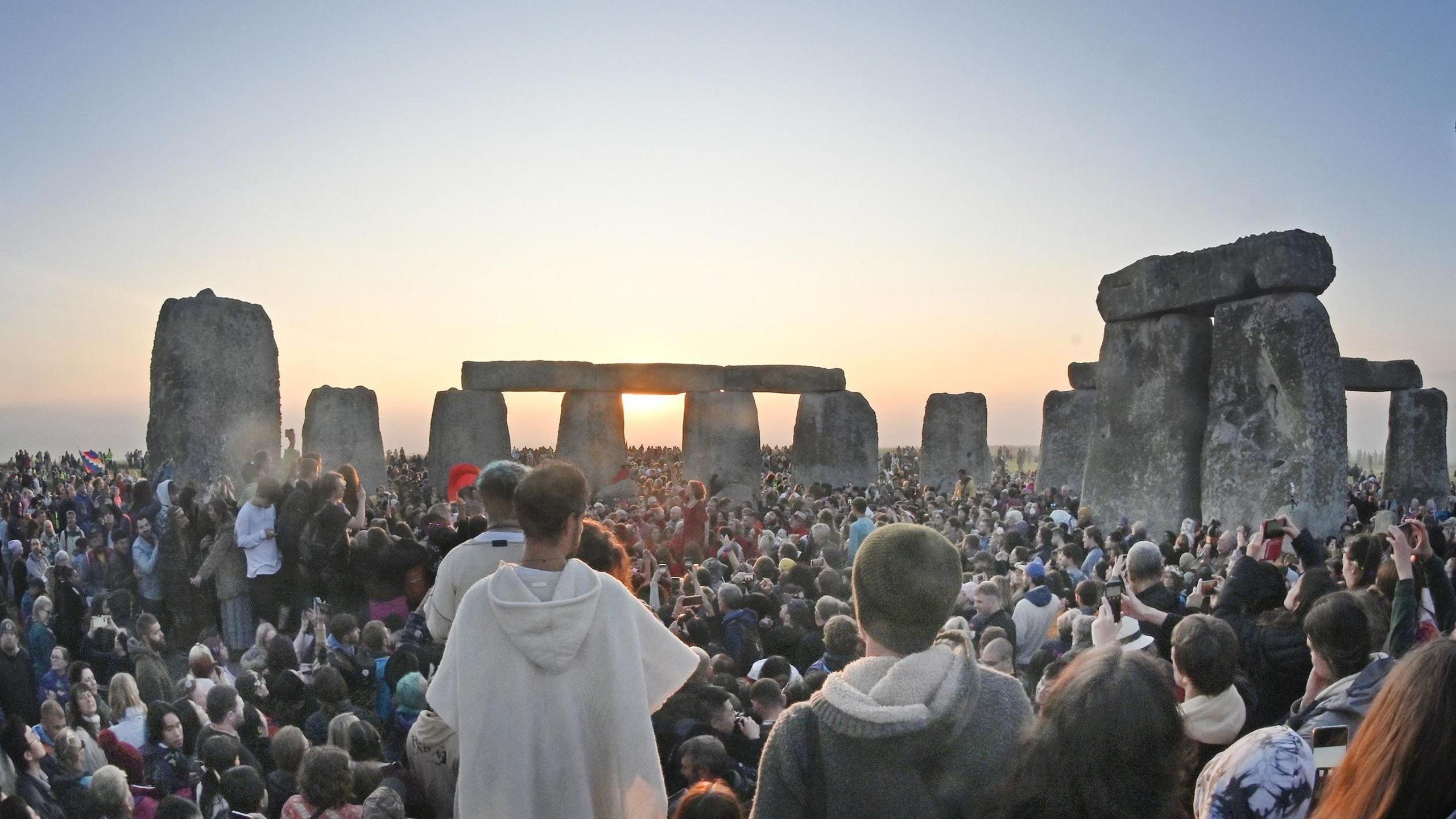Big crowds around Stonehenge's famous big stones. One person is a wearing a white coat and stood slightly higher than the crowd