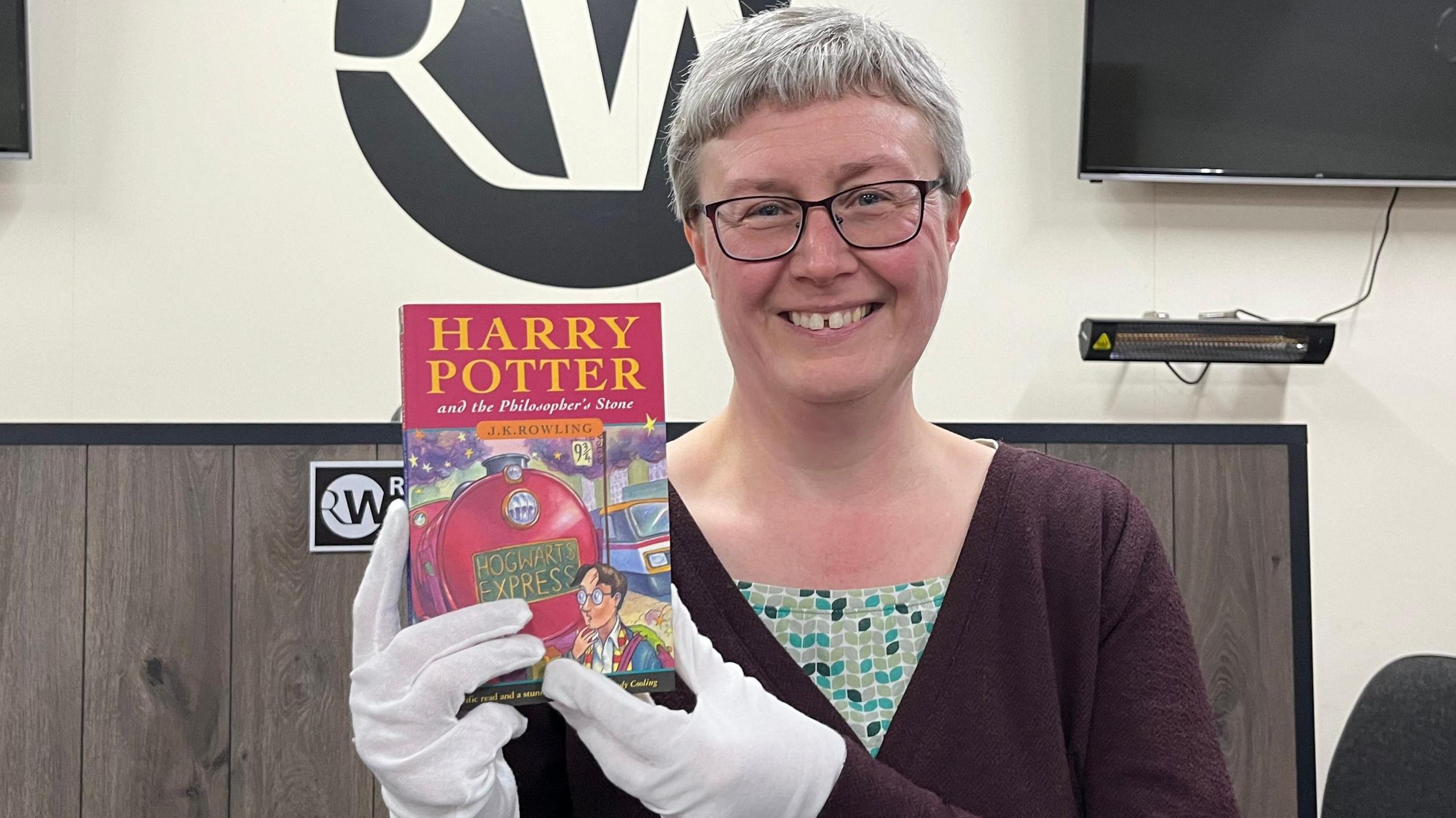 Jane Thompson-Webb, with short greying hair, holding the Harry Potter book. She is stood in front of a Richard Winterton auctioneers sign and wearing white gloves and a maroon top. 