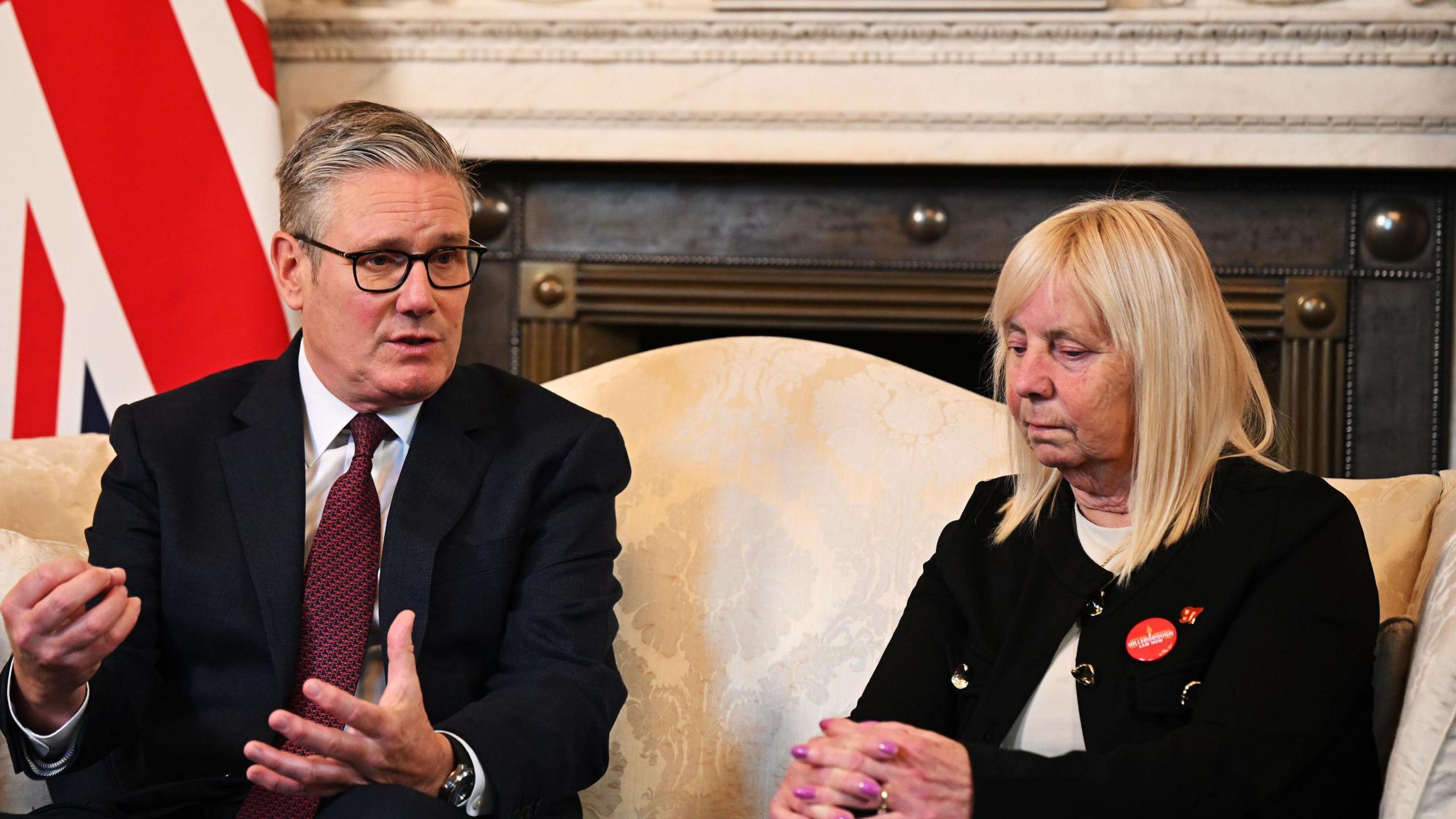Prime Minister Sir Keir Starmer speaks with Margaret Aspinall during a meeting in 10 Downing Street. They are sitting on a cream patterned sofa with a fireplace in the background and cup and saucers on a coffee table. They are both wearing dark suits.