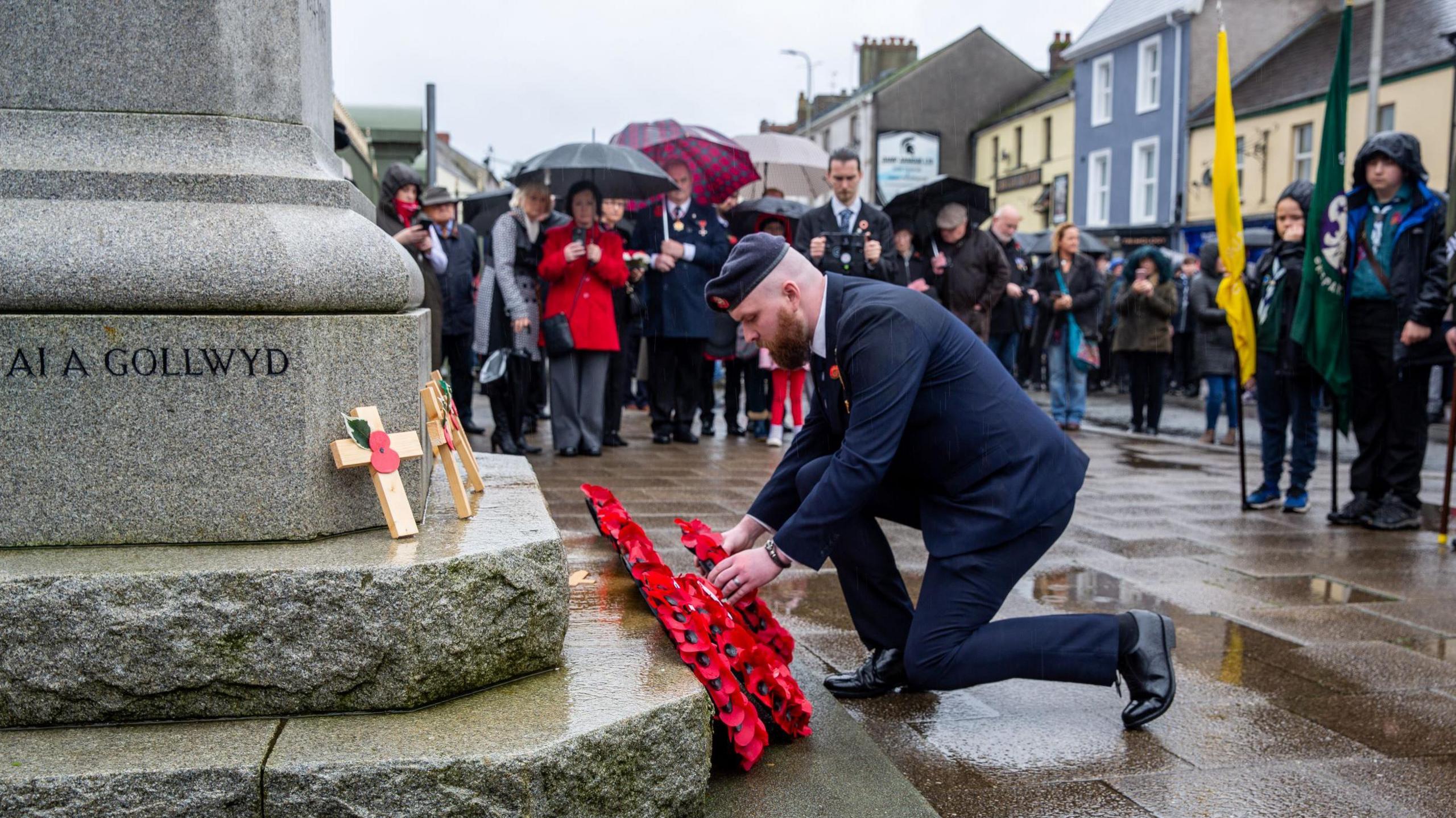 Photograph of a man kneeling on one knee to lay a poppy wreath.