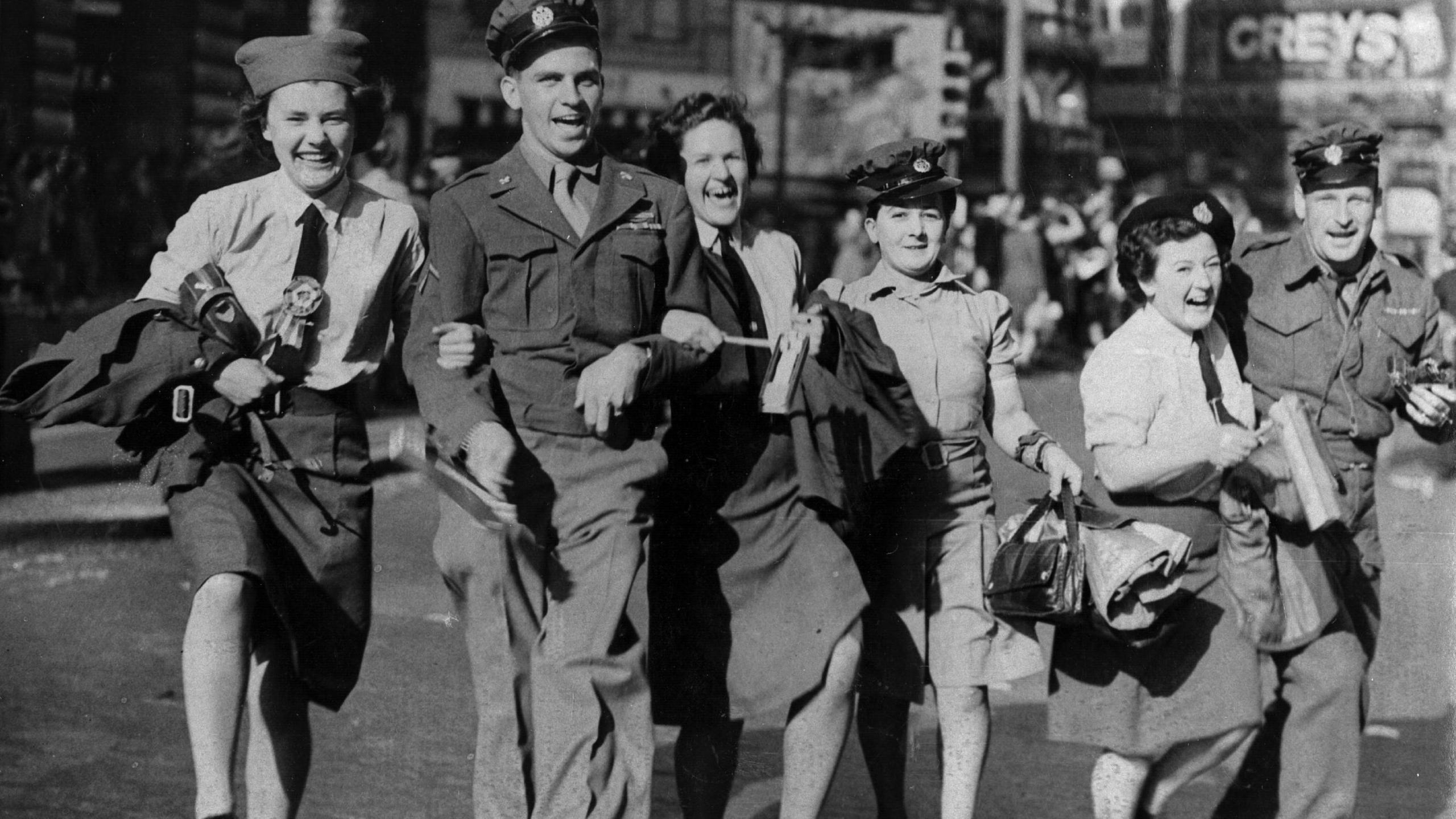 A black and white photo of a group of men and women in service uniforms.  The women wear light-coloured shirts, black ties and dark skirts, while the men wear dark grey jackets and light grey trousers. The women are wearing berets and the men are wearing dark caps. They have linked arms and are smiling as they walk down a wide road.