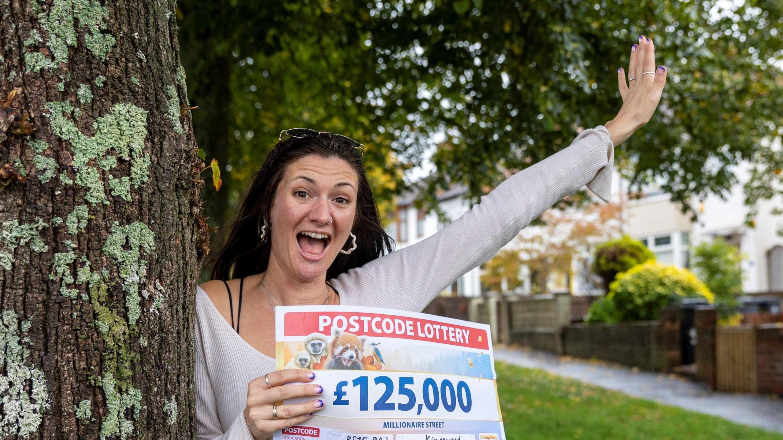 Gemma Marson-May standing by a tree on her street. She is holding the cheque from the Postcode Lottery and has her arm in the air in triumph. Her mouth is wide open with excitement.