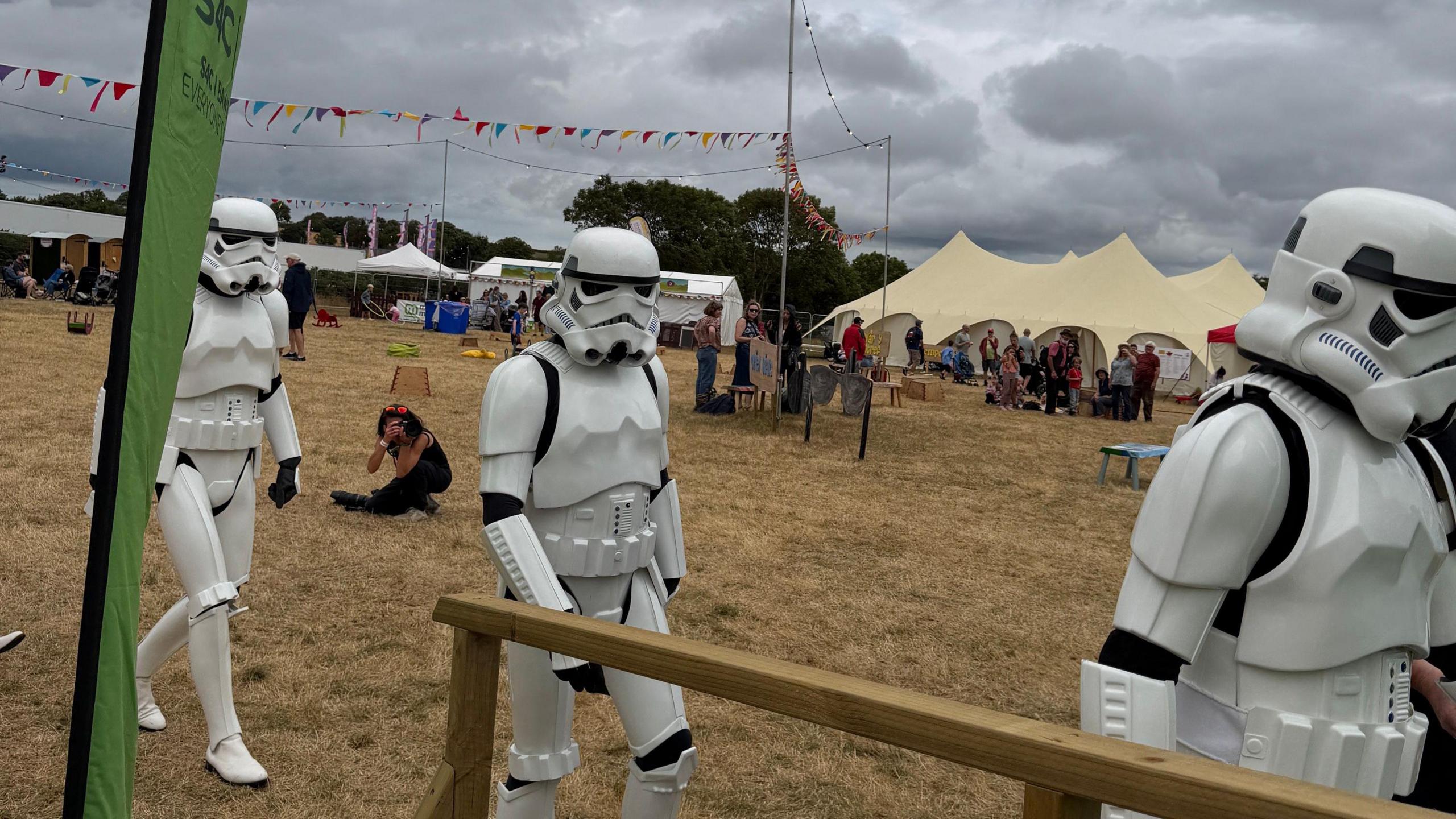 Three Stormtroopers at the National Eisteddfod, walking in a single file line. Tents can be seen in the background on the field where the festival is hosted.