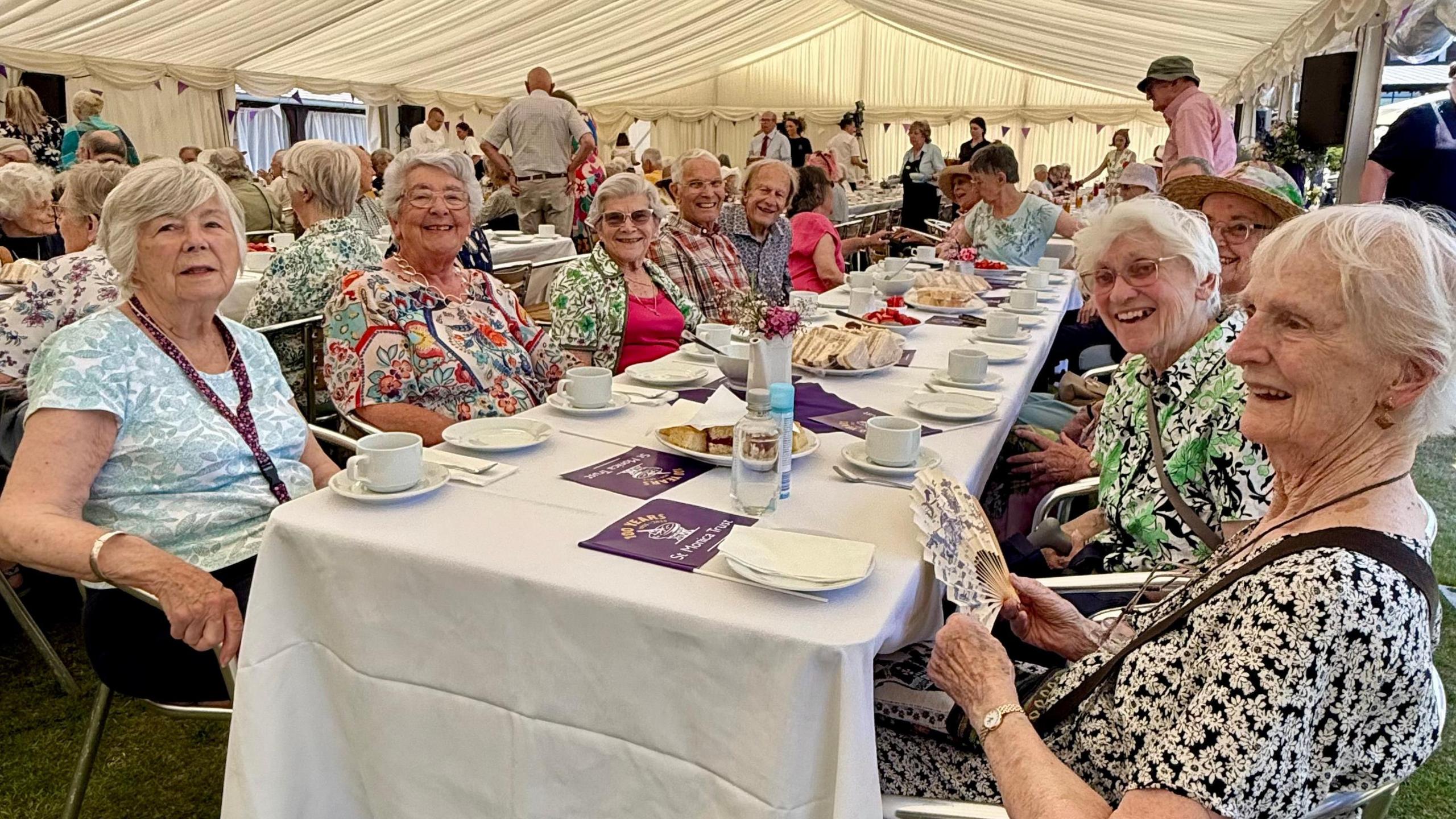 Residents of St Monica Trust villages and care homes gather for an afternoon tea. Several men and women of an older age range sit at a table, with sandwiches and cakes on it.