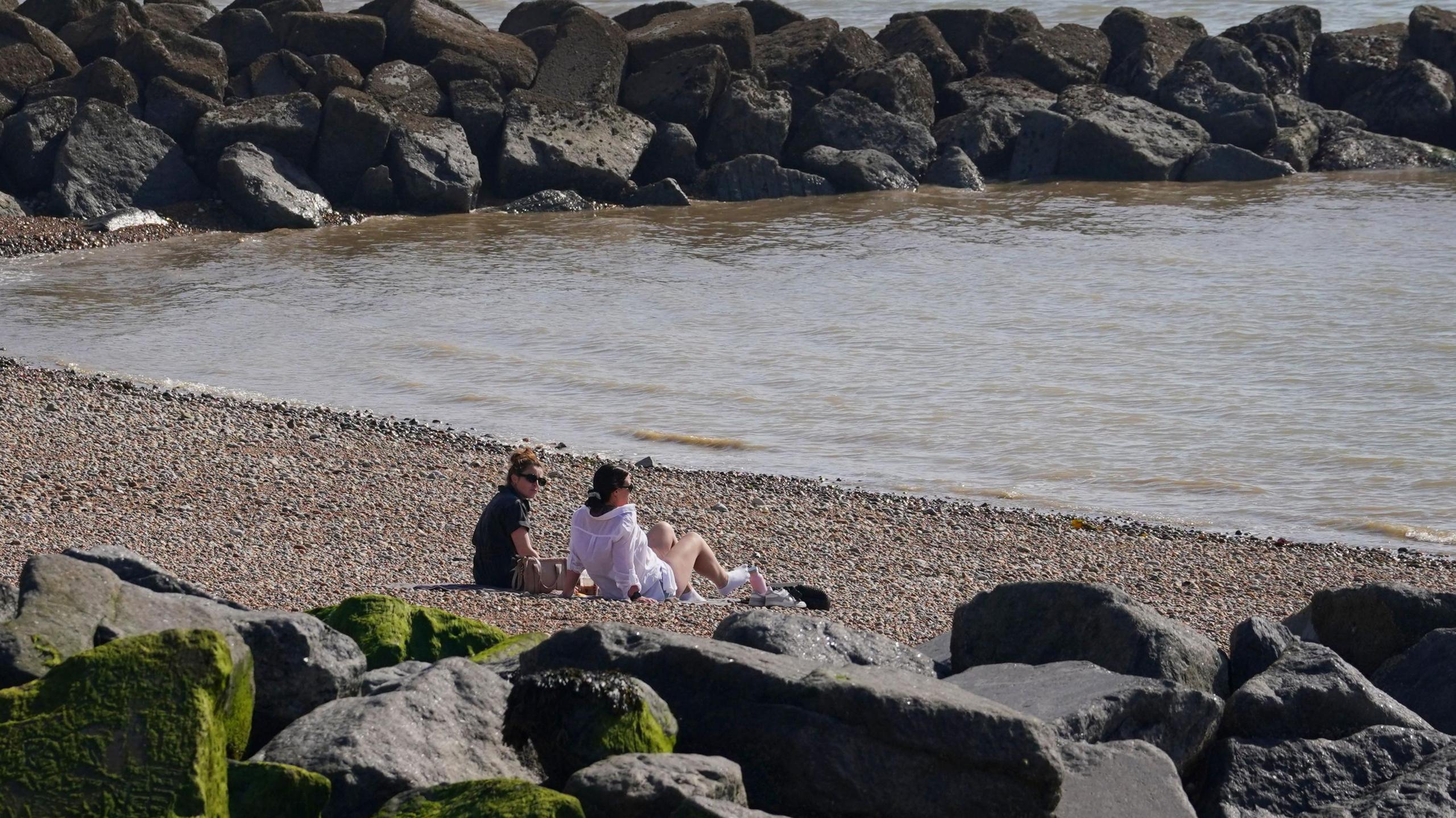 Two women relax in the sunshine on a beach. It is a rocky beach and there are large grey boulders surrounding the women.