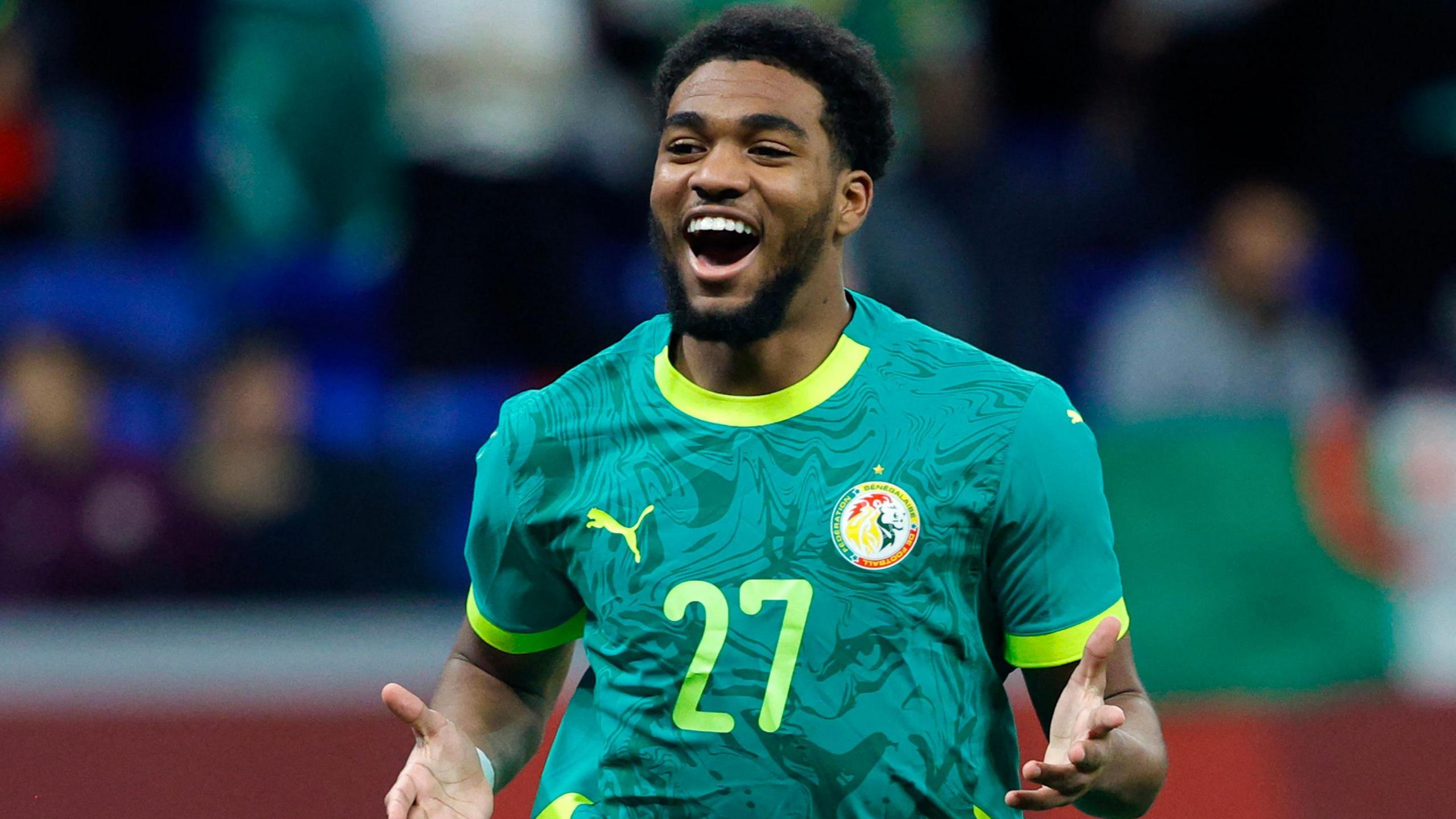 Ibrahim Mbaye, wearing a green Senegal shirt with yellow trim and a number 27 on the chest, smiles broadly as he celebrates a goal