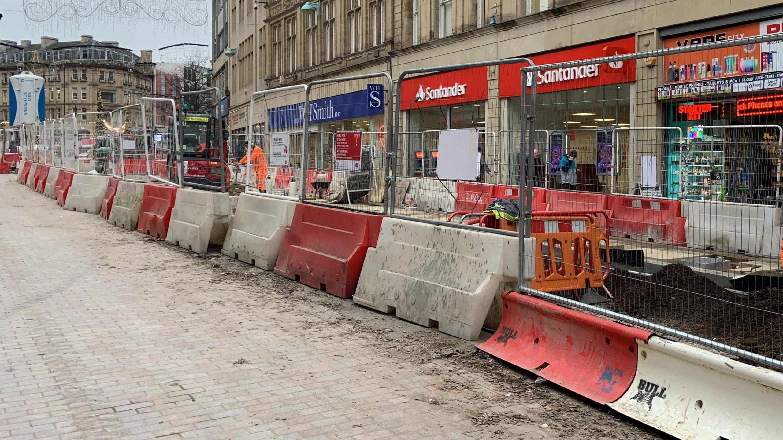 Photo shows Fargate, a shopping parade in Sheffield city centre. A line of heavy red and white plastic blocks and high fences runs the length of the precinct, in front of several High Street shops