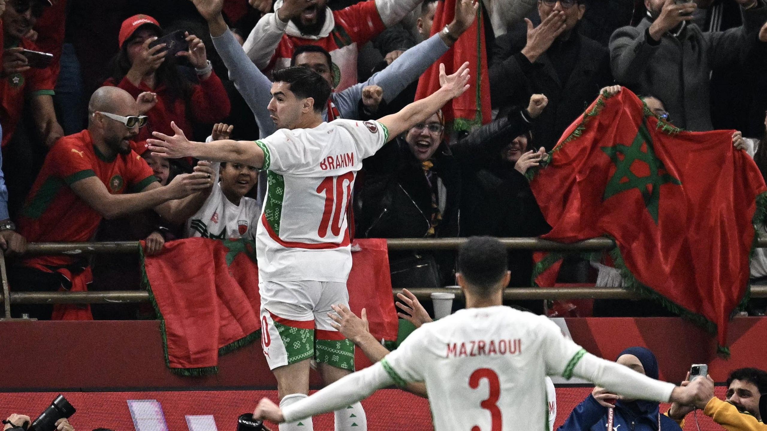 Brahim Diaz of Morocco is shown from behind as he stands on a advertising hoarding at the Stade Prince Moulay Abdallah during Afcon 2025 to take the applause of the passionate Moroccan crowd facing him. Diaz is in white kit with green and red trim and the his name and the number 10 in red on his back. He has his arms spread wide to take the applause and is turning his head slightly to his left. In front of him the fans raise their arms and wave Moroccan flags, while team-mate Noussair Mazraoui can be seen running towards Diaz, also with his arms spread wide