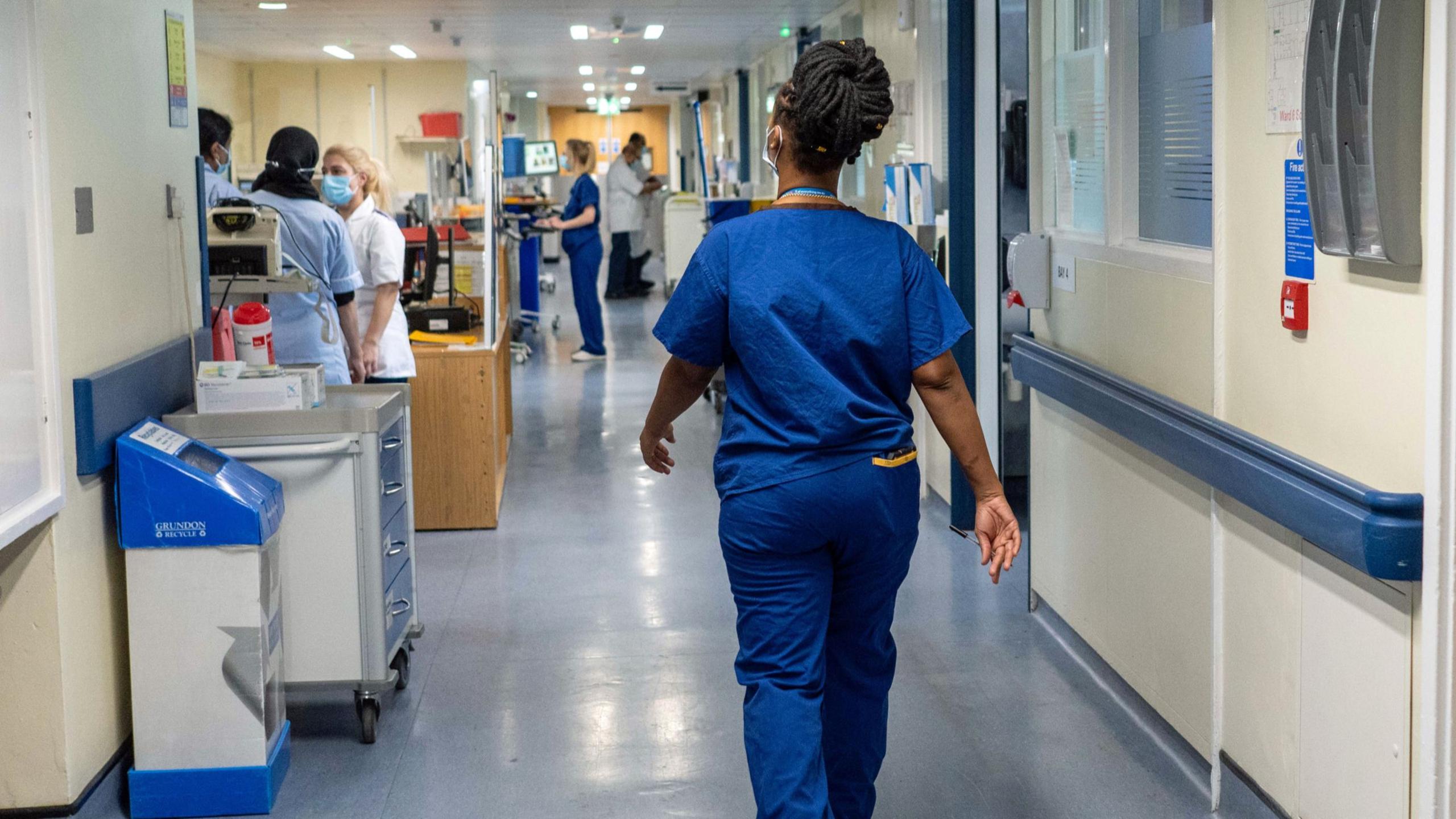 A doctor wearing blue scrubs walks down a busy hospital corridor