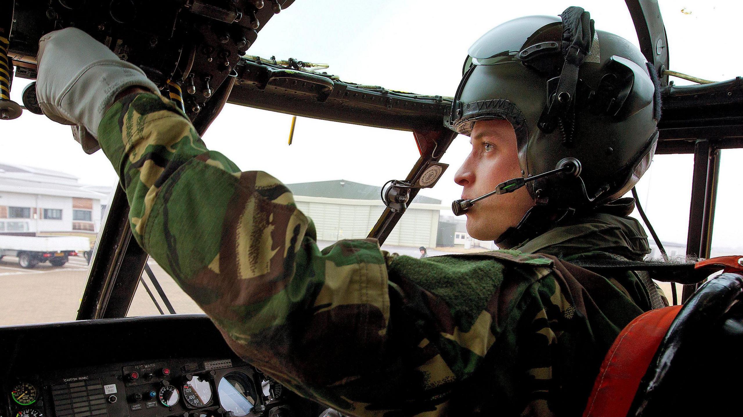 Prince William in the cockpit of an aircraft. Is is wearing a helmet and a green and brown military camouflage flying suit .