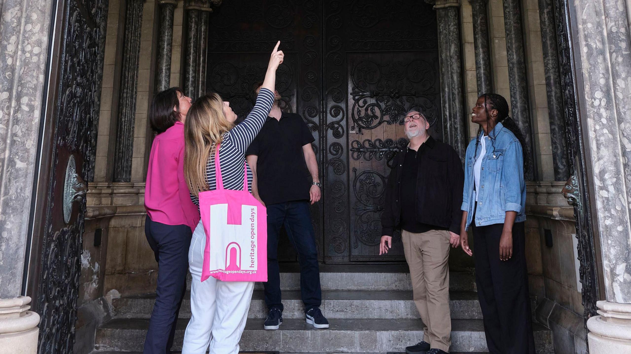 5 people stand under the vaulted stone arch of a church. The woman in the foreground is holding a bright pink bag with a "H" on it standing for Heritage Open Days. 