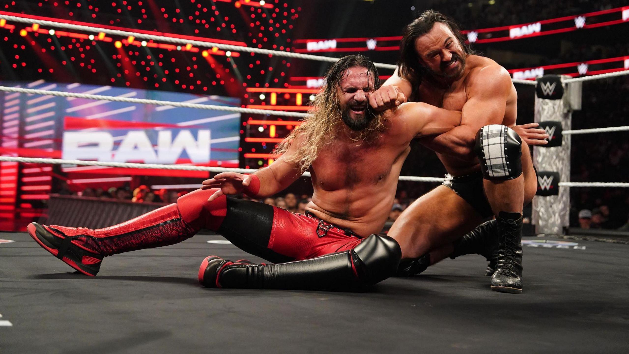 Drew McIntyre (black trunks and boots) holds an armbar on Seth Rollins (red and black trousers and boots). Both men are grimacing with effort. 