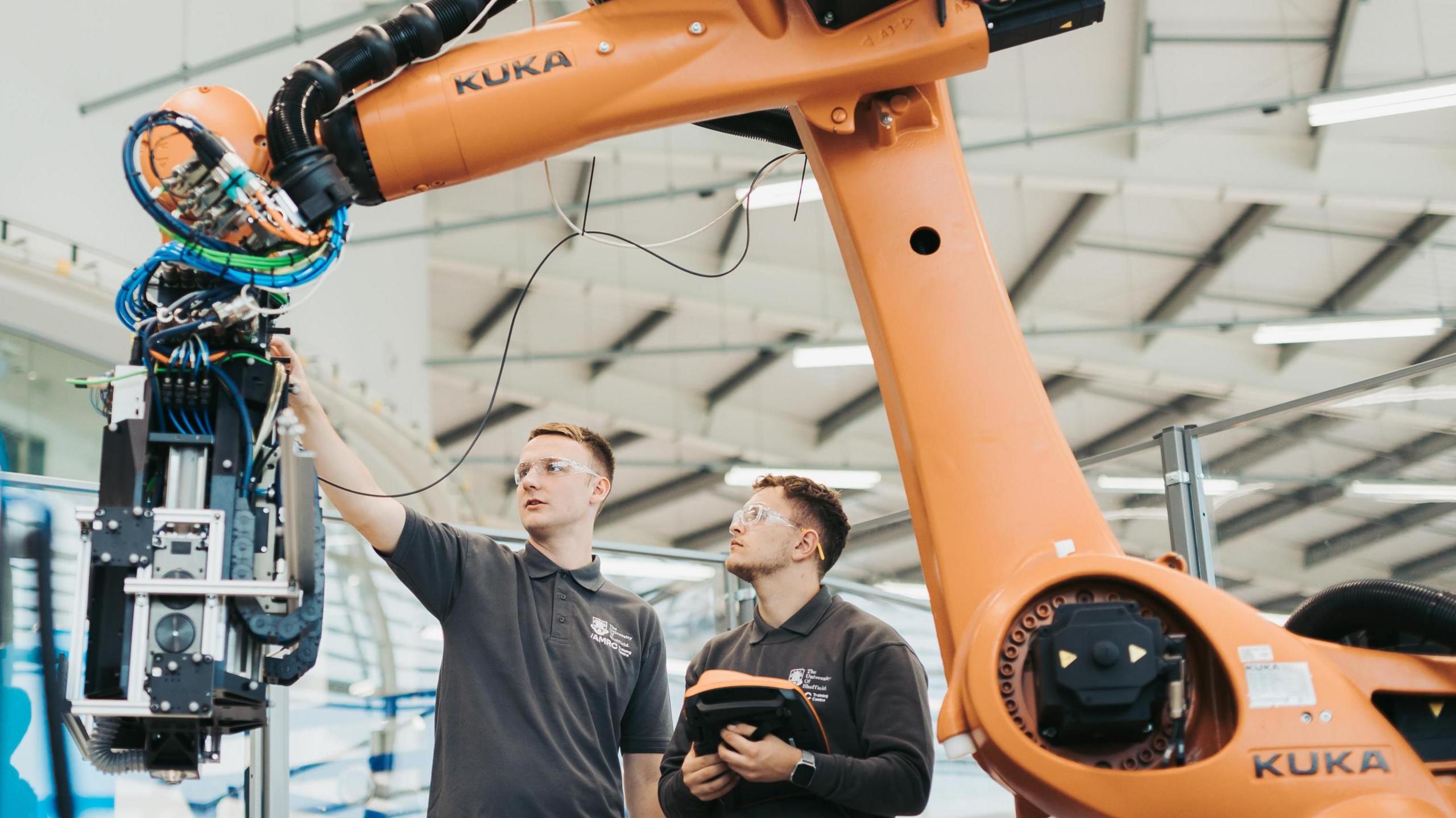 Two men wearing clear safety goggles are stood looking at a yellow robotic arm