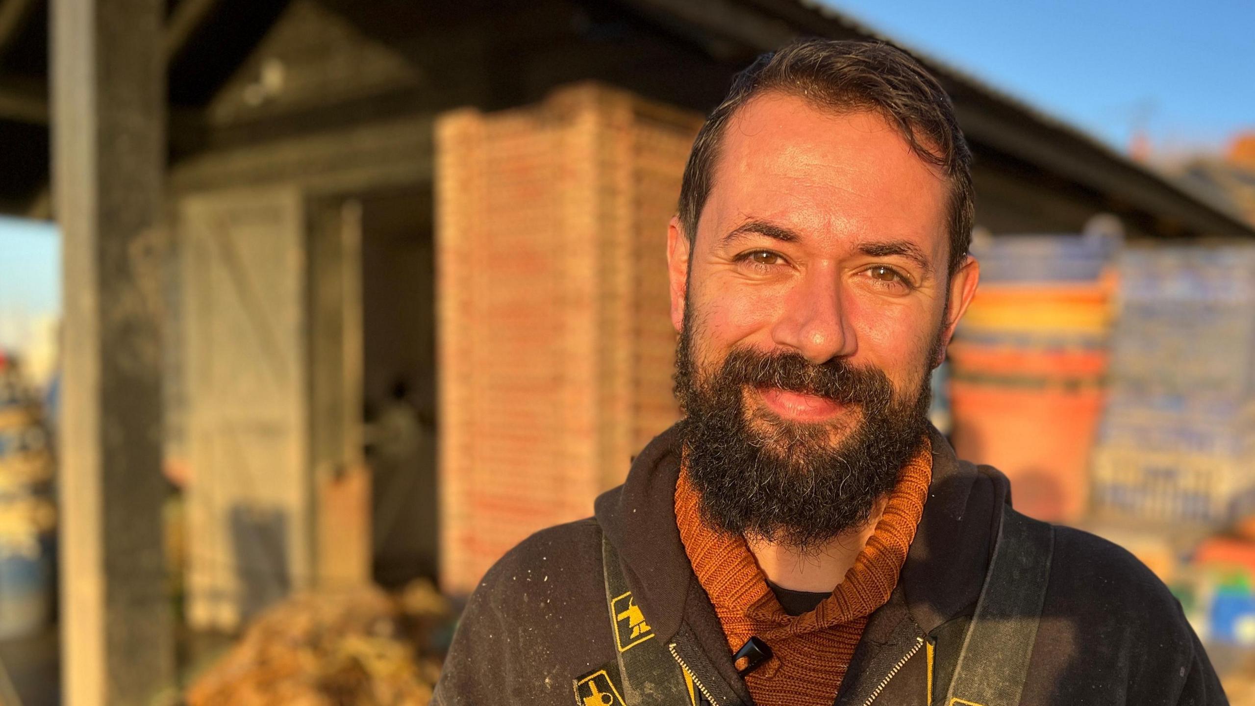 Tom Haward, a man in his early forties with a thick beard, smiling directly at the camera. He is standing in front of his oyster shed on Mersea Island in Essex.