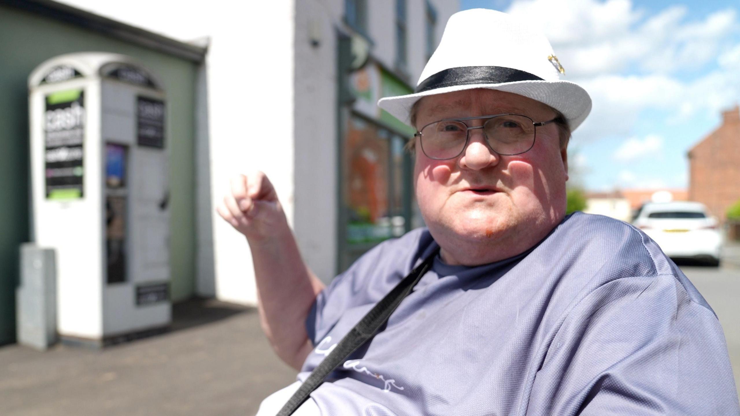 66-year-old Nigel Charlton in front of a white phone box, with cash machine stickers on it. Nigel has rosy cheeks and is looking into the camera, wearing thin-framed grey glasses with clear lenses, a purple t-shirt and a white panama hat with a black ribbon around it. His right arm is raised.