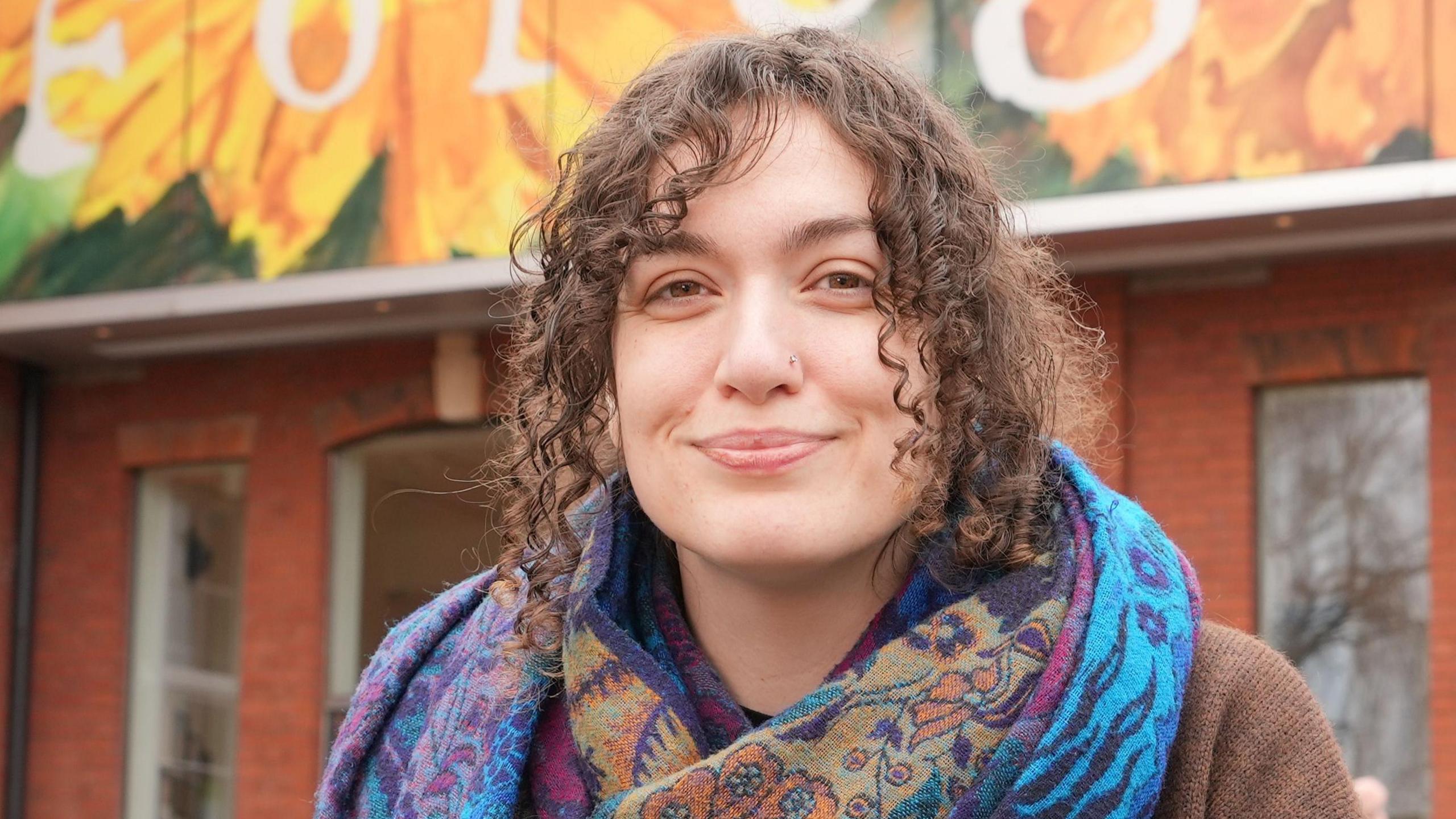 Steph smiles into the camera. She has dark brown, curly hair that is tied up, and is wearing a blue patterned scarf and a brown cardigan.