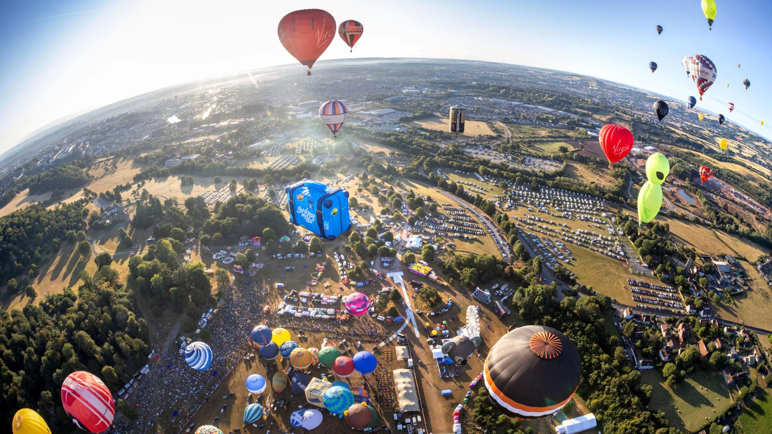 An aerial view of the entire Bristol Balloon Fiesta site showing parking areas, the crowd and dozens of balloons either in flight or about to take off