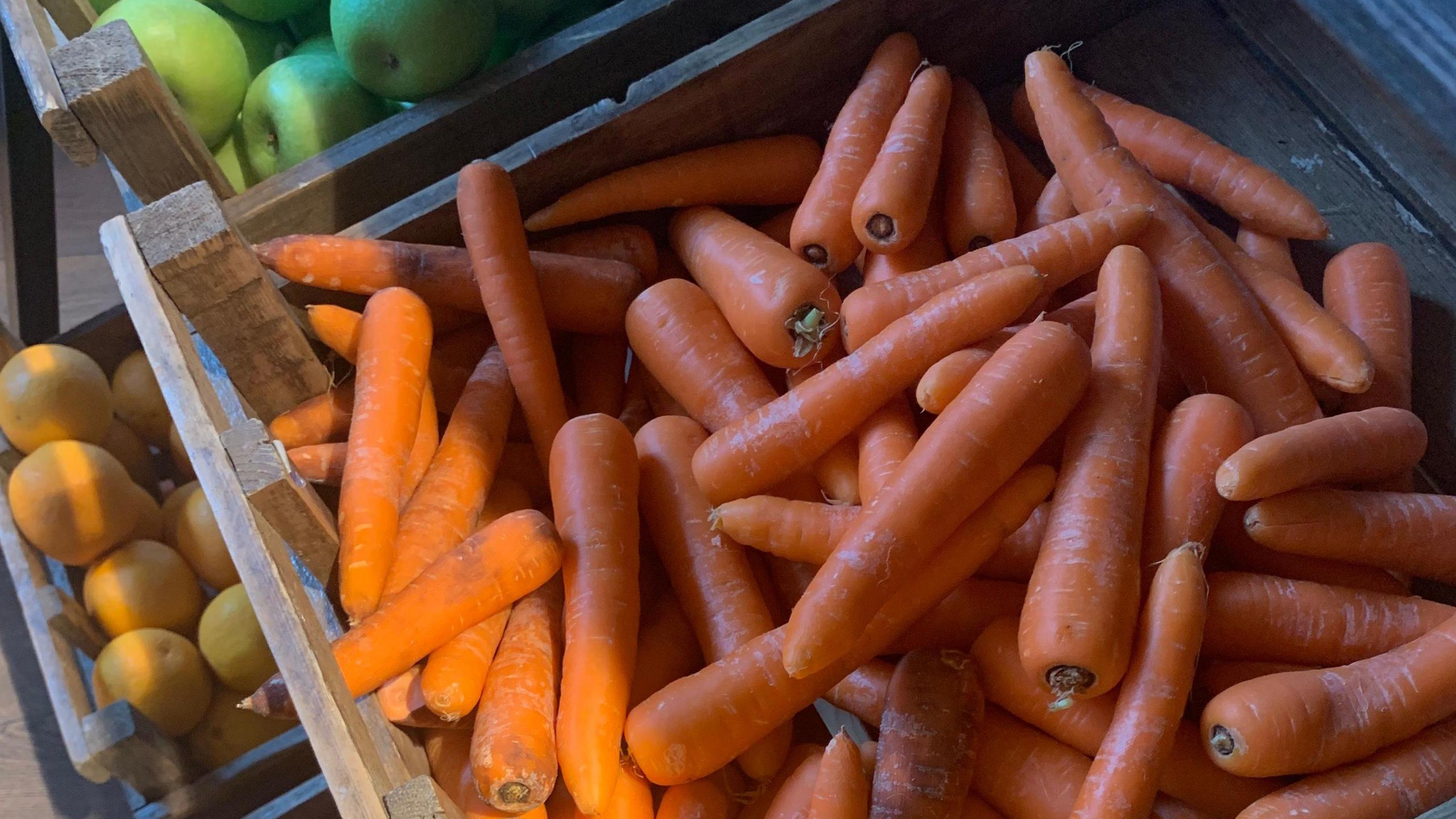 Wooden boxes filled with orange carrots, green apples and oranges. A streak of sunlight illumines the edge of the boxes.