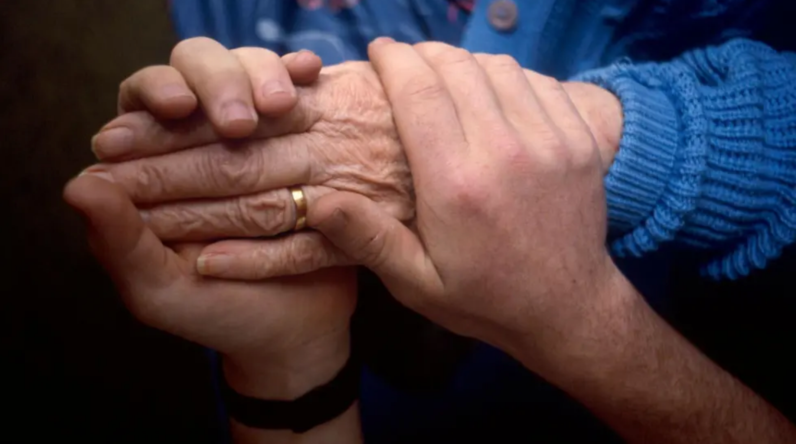 A younger person holds and older person's hand. The older person has a glue cardigan on and a gold wedding band.