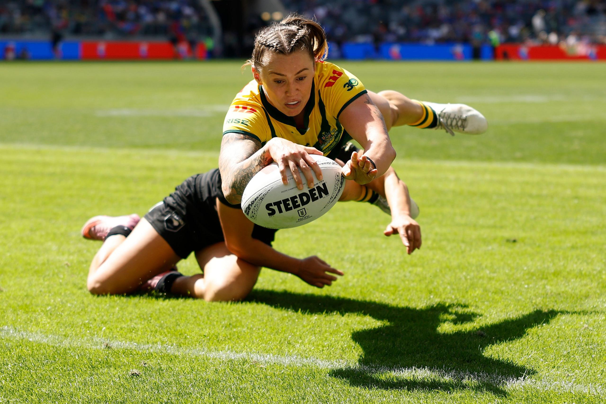 Rugby player in a yellow and green jersey diving forward while holding a Rugby ball, tackled by an opponent in black on a grassy field, with stadium seating and colorful advertising boards visible in the background under bright daylight.