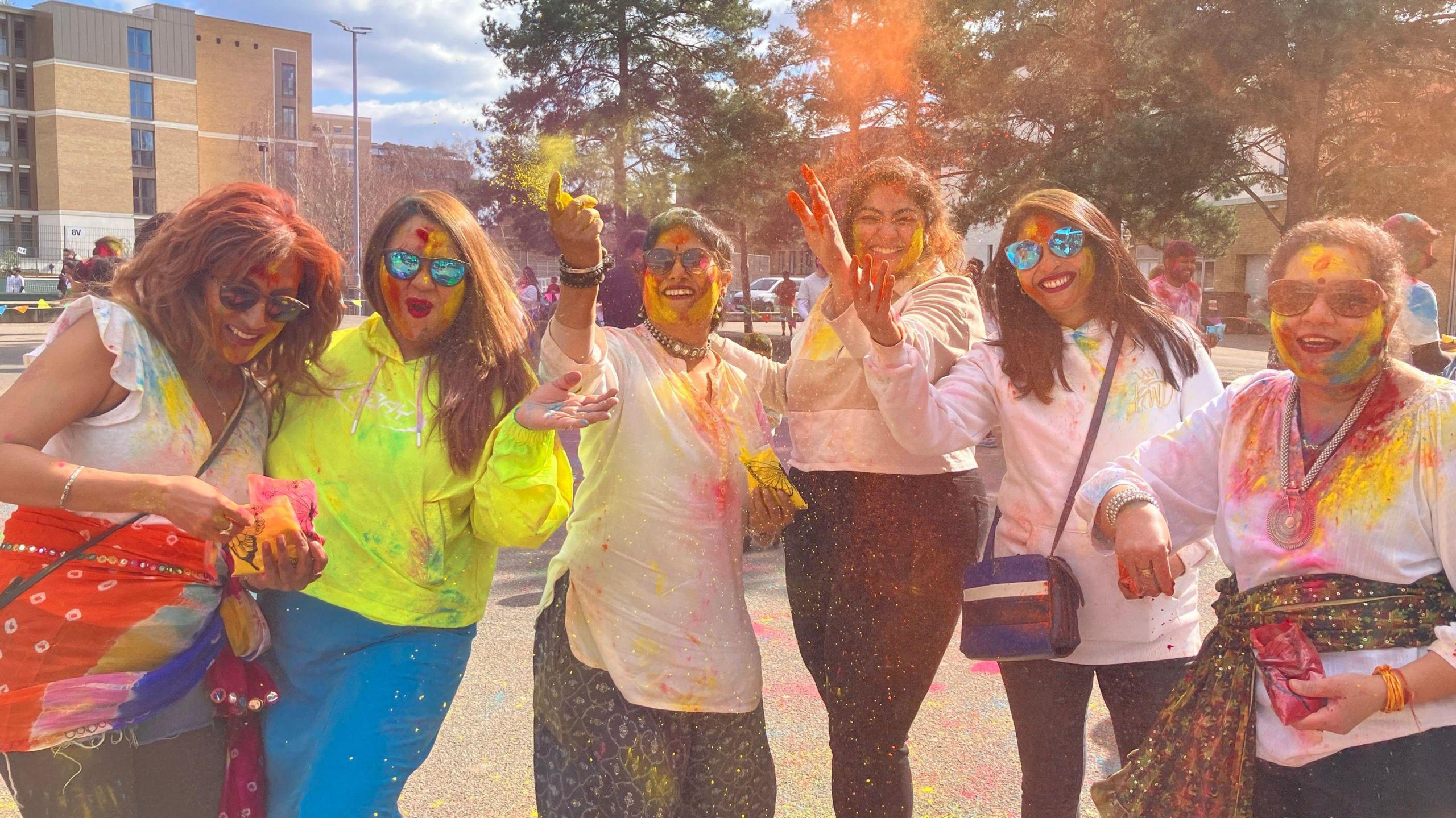 A group of five women laugh and pose for the camera as they throw colourful powder over each other. 