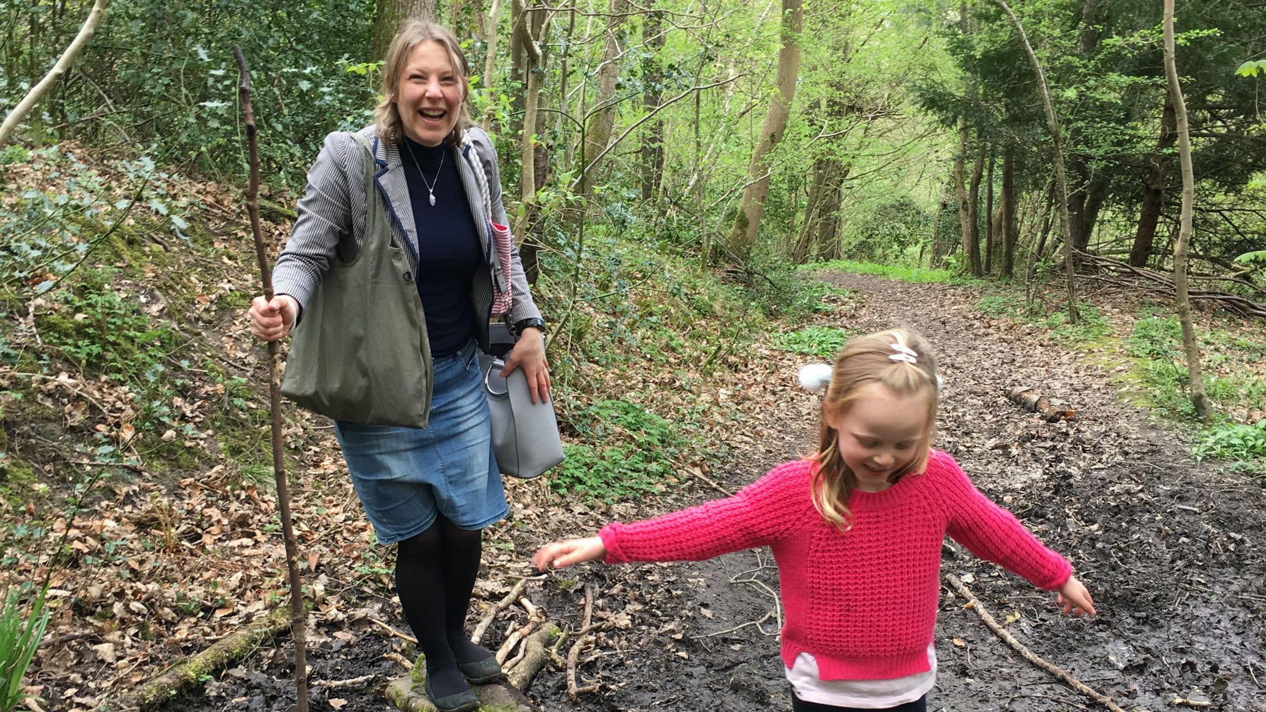 A smiling Mrs Clark, wearing a grey jacket, black top and blue denim skirt, holds a large stick as she walks in a woodland area with her young daughter who wears a red jumper.