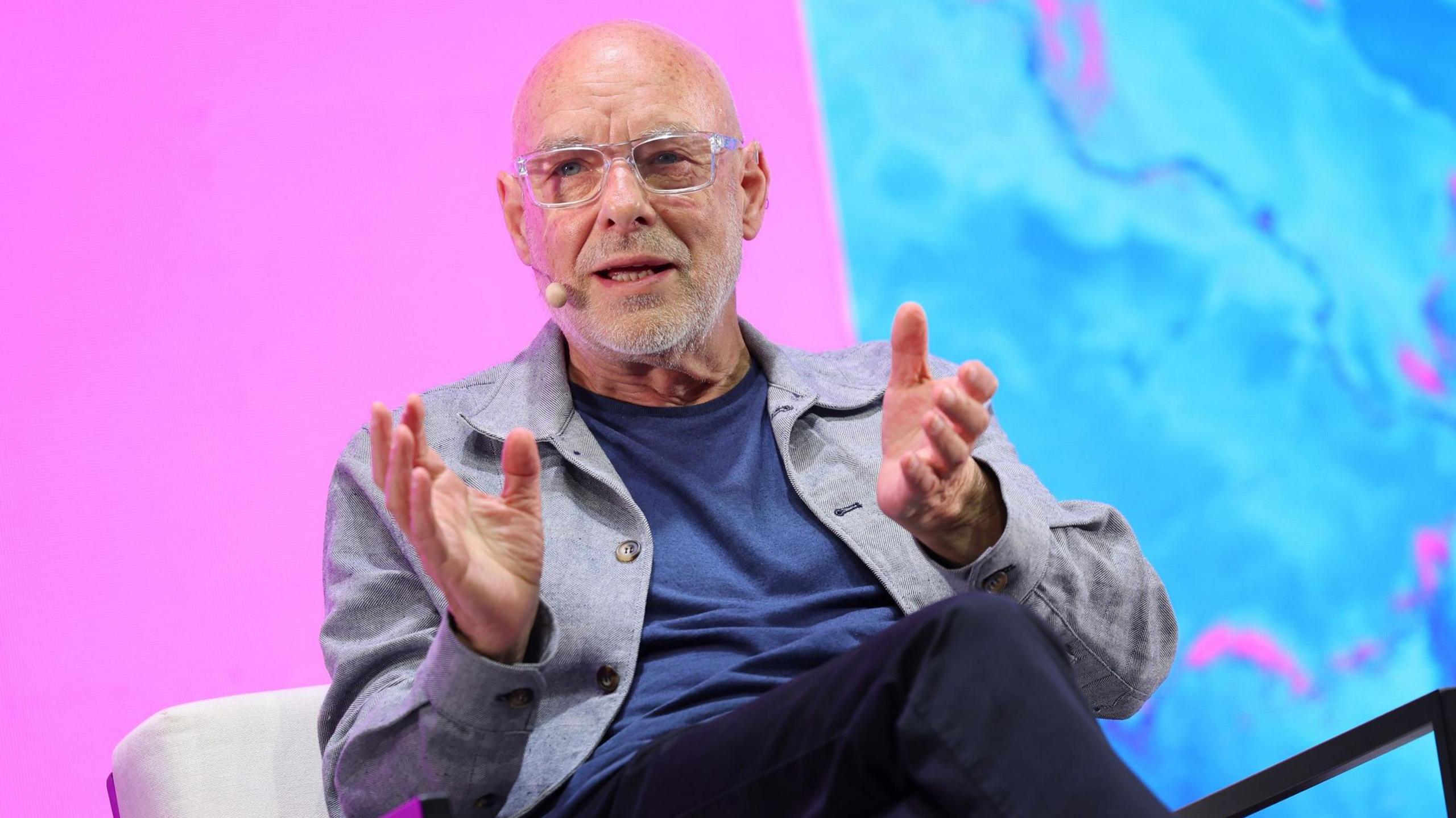 Brian Eno sitting on a chair talking at a conference. He wears a grey blazer over a blue t-shirt. The background screen is bright pink and blue.