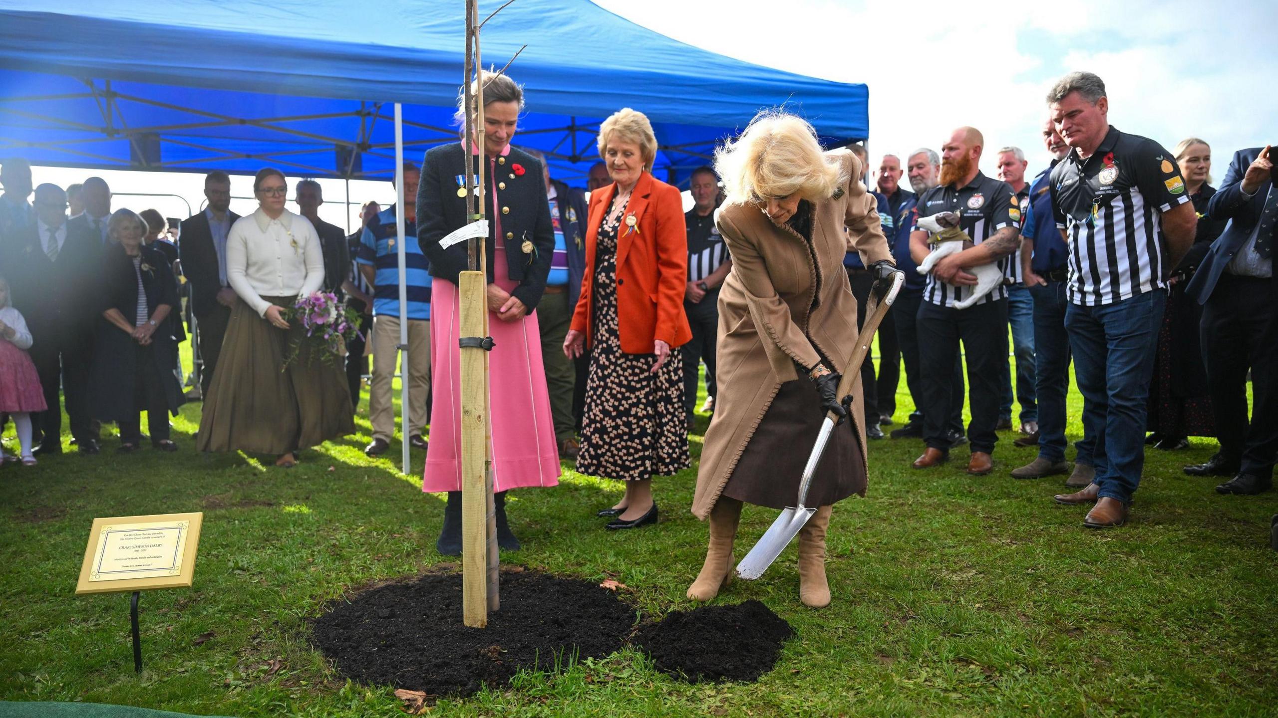 Queen Camilla, wearing a brown coat and carrying spade, plants a tree in memory of Craig Dalby, an ex close protection officer, during her visit to Bromham Community Hub. Two women are stood close by wearing pink and red winter coats.