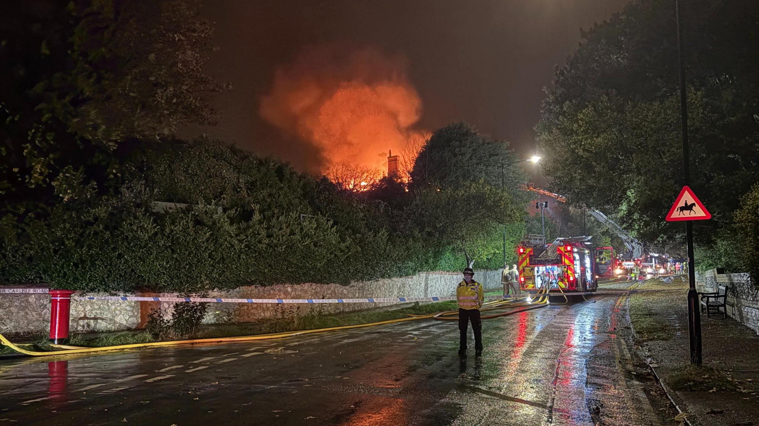 A police officer guards a sealed-off road, with a fire engine behind it and, in the distance on a hill, a large building well on fire.