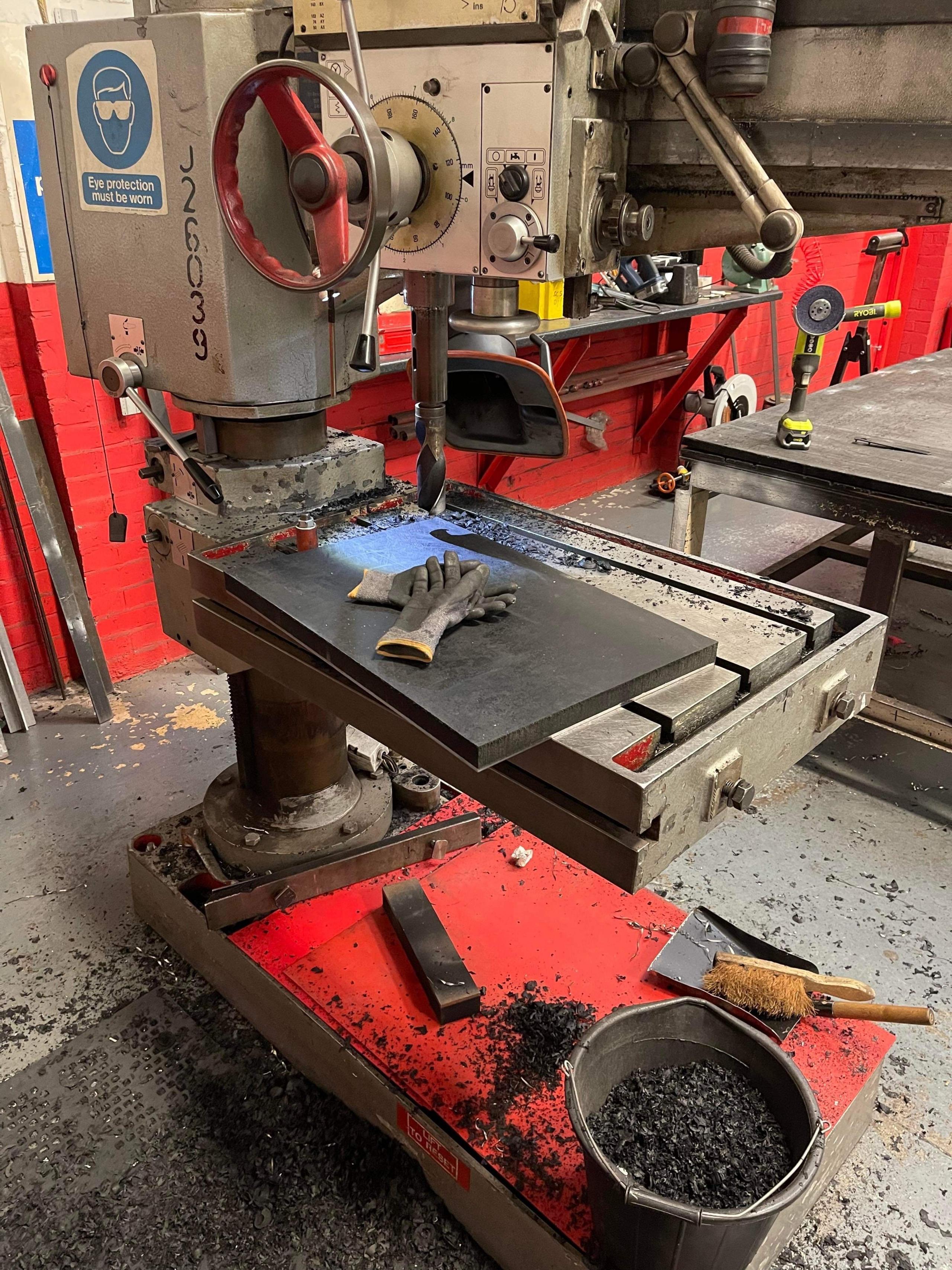 A silver industrial drill, with a red wheel on the side and a white and blue sticker warning that workers must wear glasses while operating. There are a pair of black gloves on the surface of the drill and a bucket of black filings on the floor