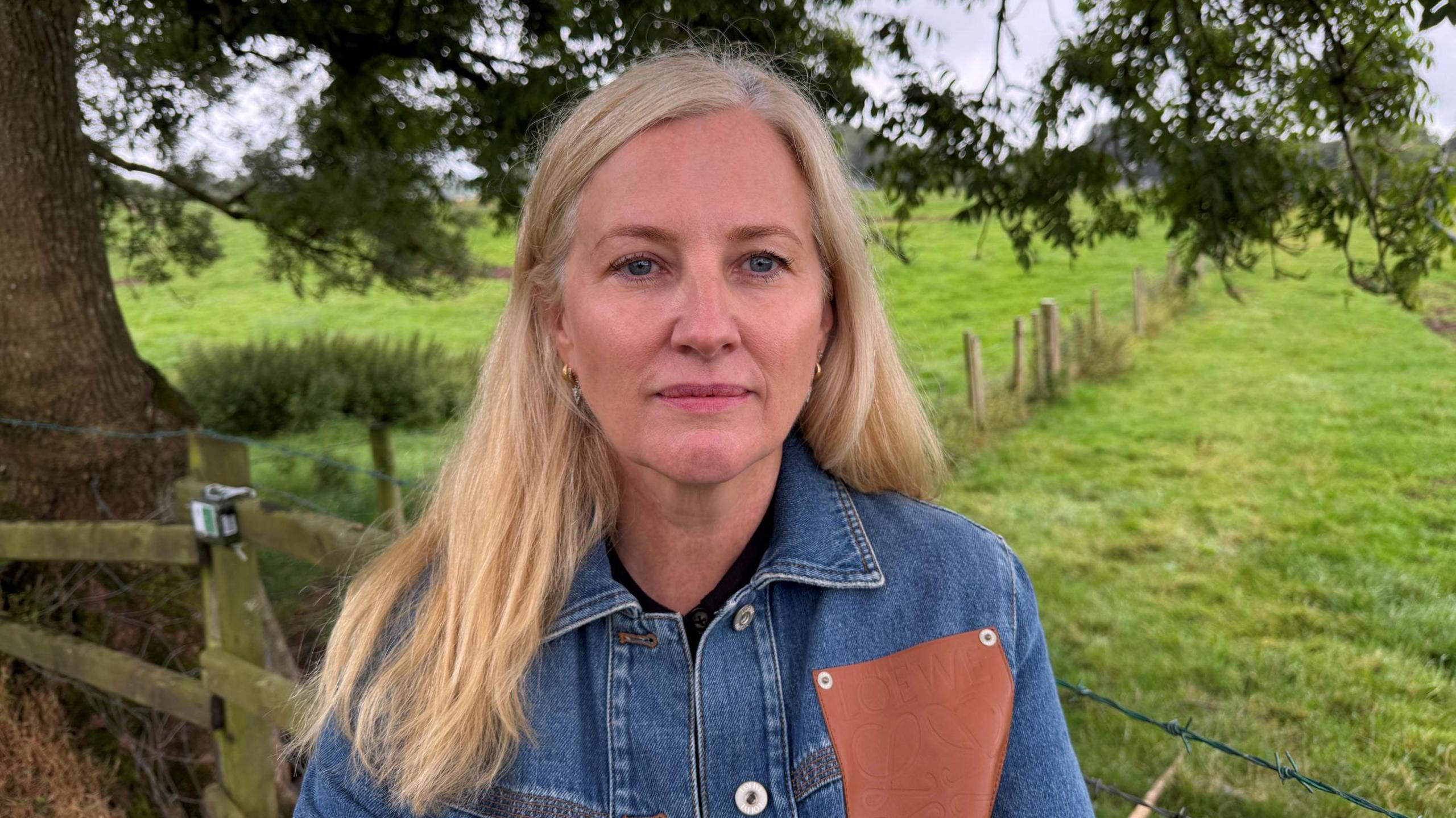 Sarah Herbison has blonde hair and a denim jacket with a brown quare on it. She is standing in front of a field with part of a tree behind her.