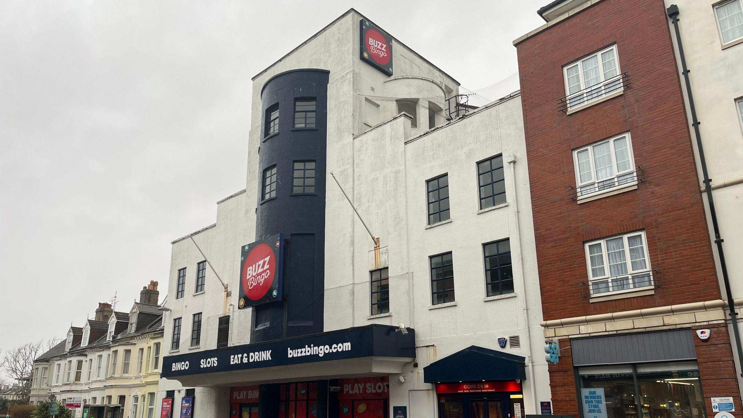 The Buzz Bingo building is an art deco style cream building which is four storeys tall. It has a large buzz bingo red sign on the front. There are cars parked outside the front