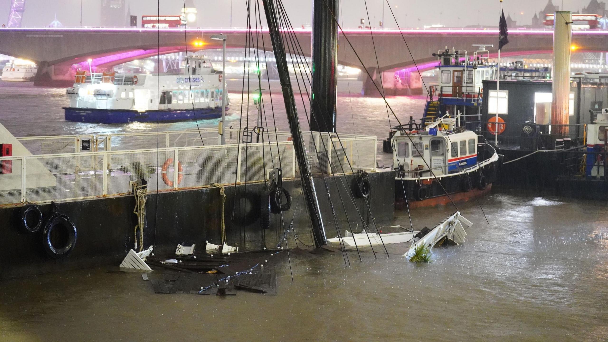 The mast of a boat protrudes from the water of the River Thames in London. The black mast sits next to the pier it was moored to. 