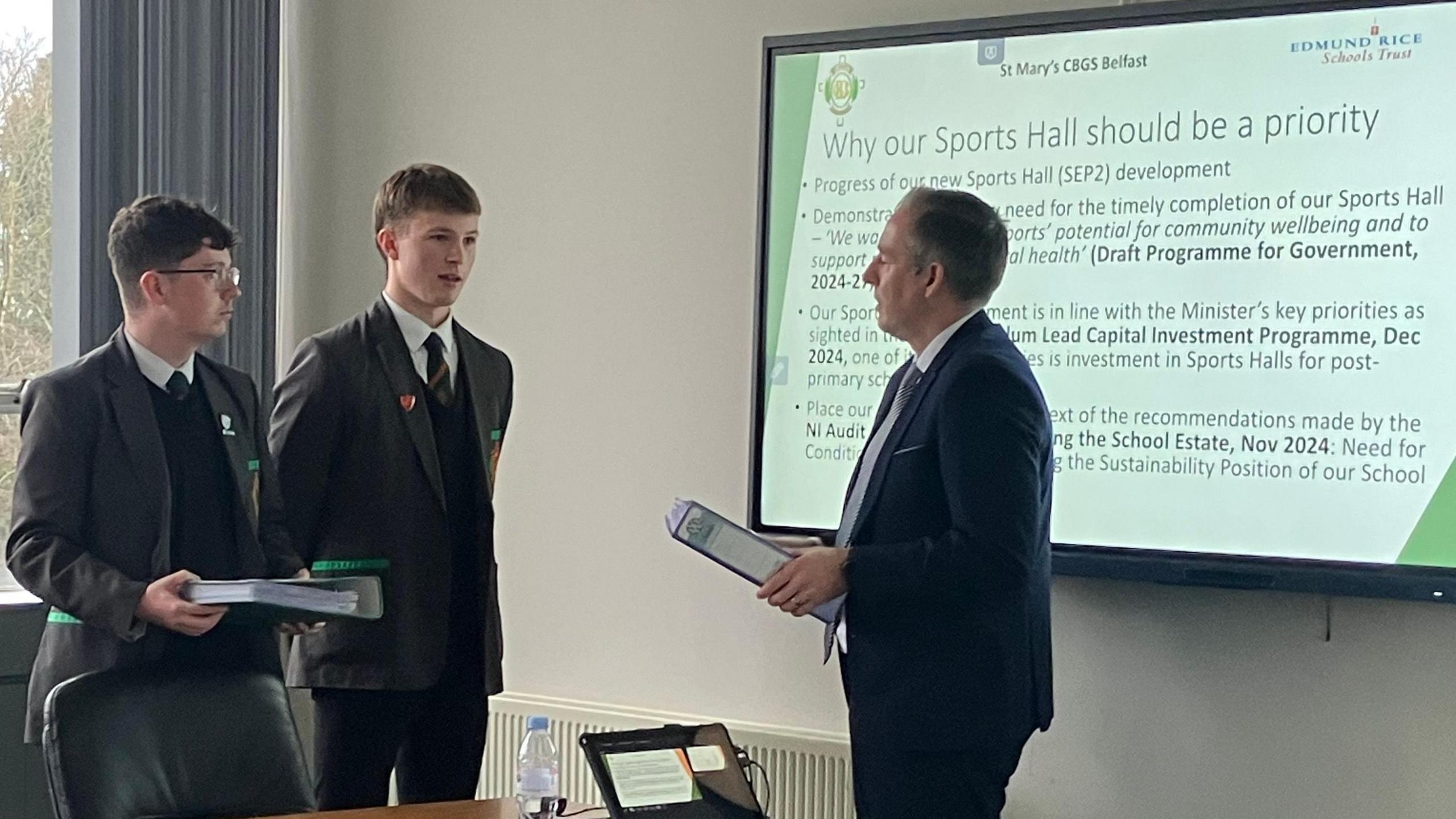 Paul Givan talking to two male pupils. The boys are wearing their black and grey school uniform, one has dark brown hair and wearing glasses and holding a folder, the other has light brown hair and has his hands behind his back. Paul Givan is holding a folder and wearing a dark blue suit. A power point presentation can be seen in the background reading "Why our sports hall should be a priority"