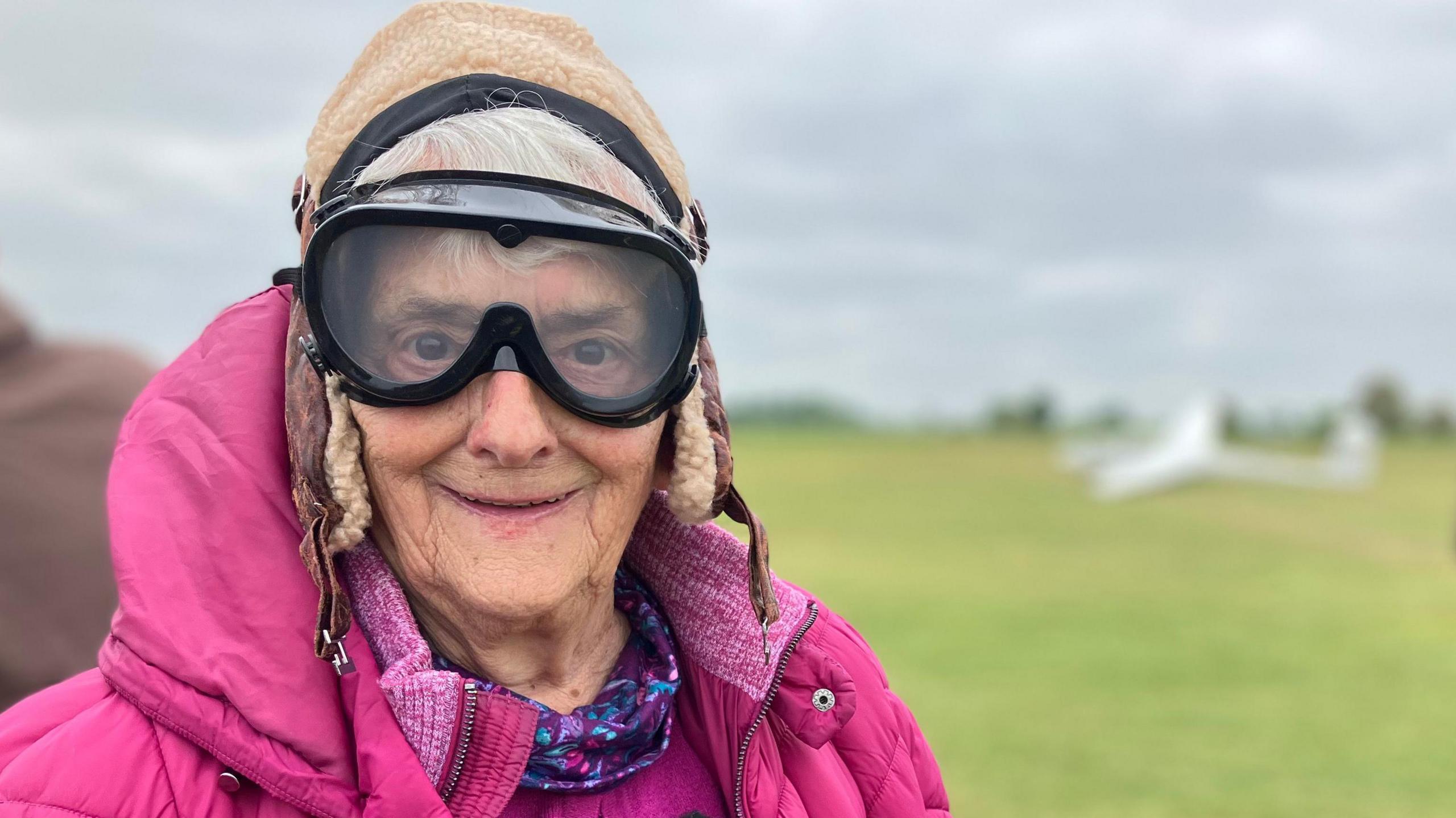 Barbara Brookes, 90, wearing black goggles and a pink coat looking at the camera with a glider in the background on a field at Husbands Bosworth Airfield, in Leicestershire