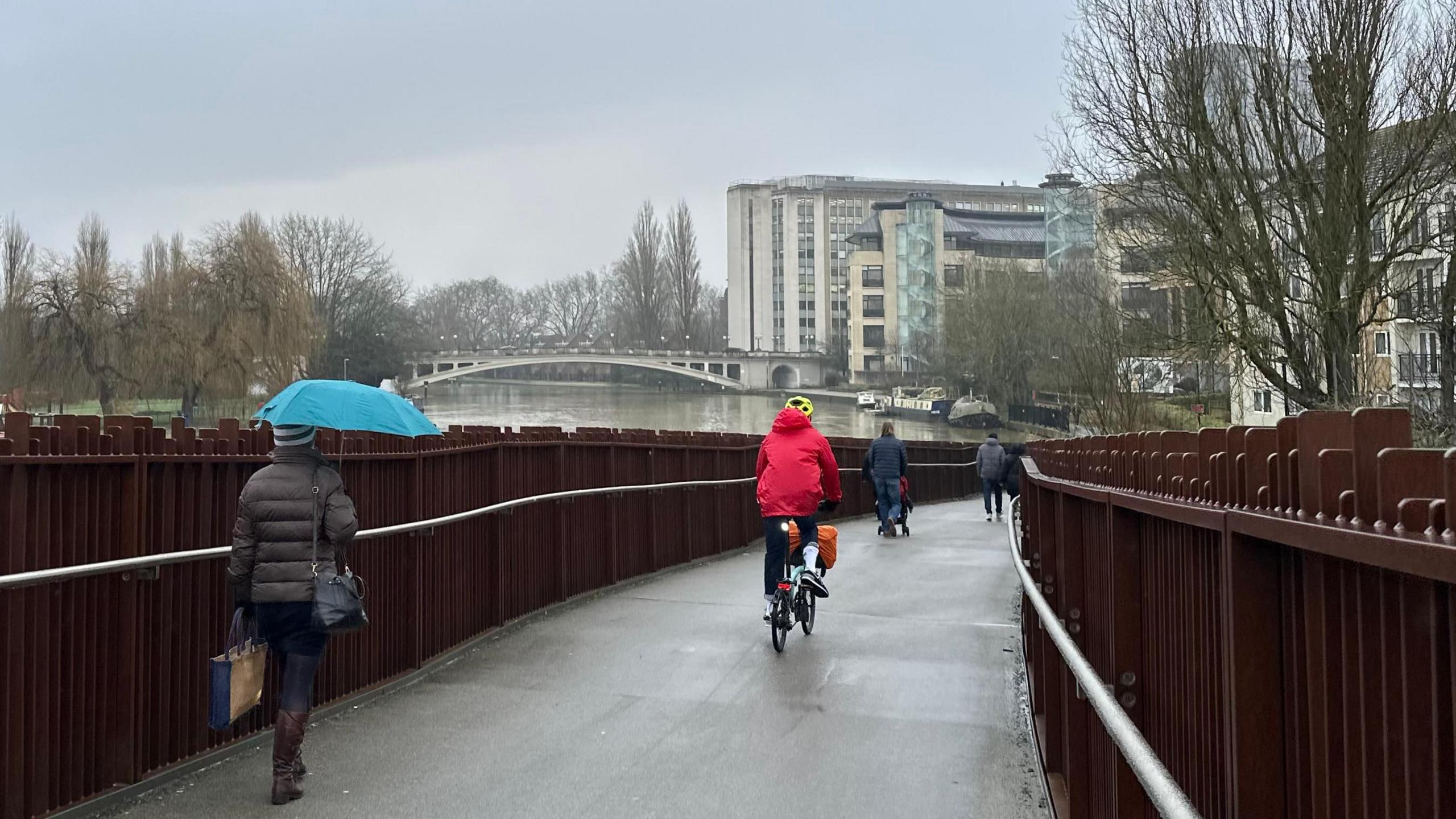 A bridge over a river with a brown fence on each side. There is a woman holding a blue umbrella and another person wearing a red coat and a yellow helmet. Over the bridge you can see the river, several high-rise buildings and a second bridge over the river in the distance. 