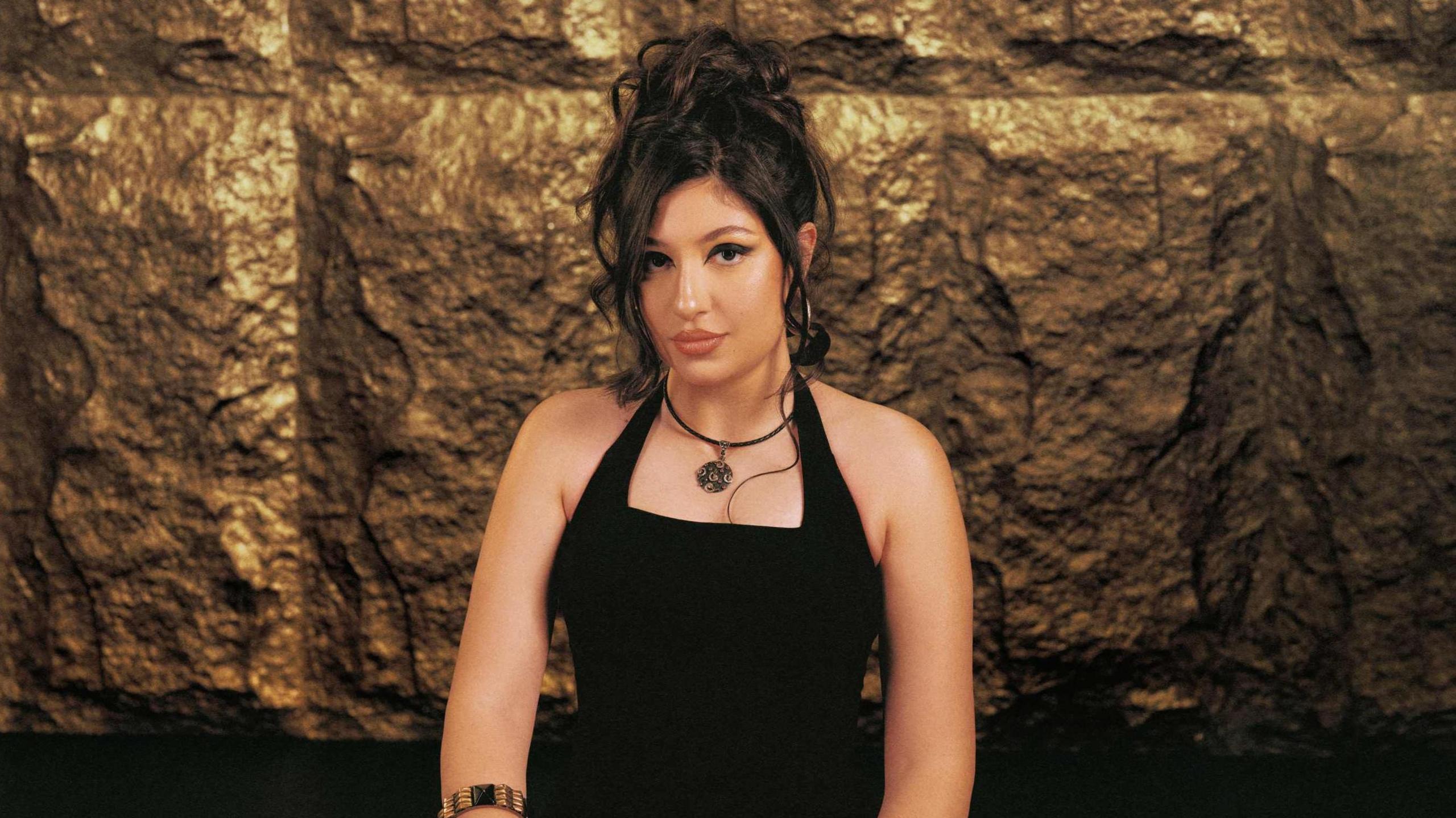 Lella Fadda wears a black dress and black necklace with pendanty. She has long black hair tied up with some of the hair framing her face