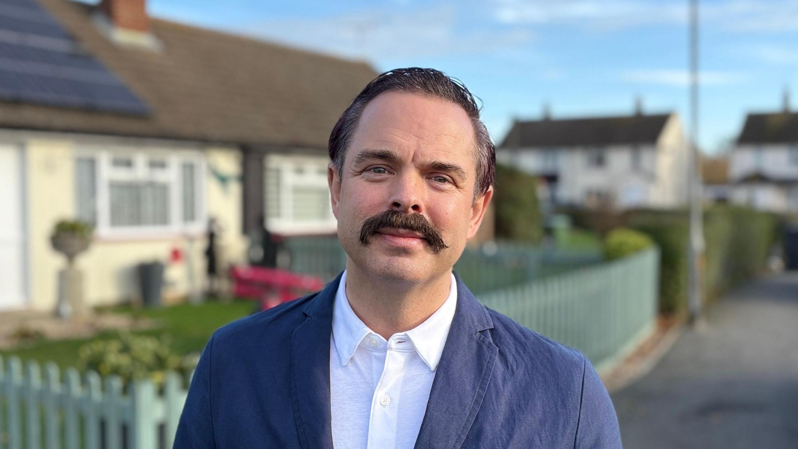 Daniel Greenwood looking directly down the camera. He has dark hair worn in a side parting and a long moustache hanging over his top lip. He is wearing a light blue jacket and white shirt, which is open at the neck. He is standing on a residential street with bungalows in the background, which are out of focus. A light green picket fence can be seen immediately behind him, running along the boundary of the nearest bungalow. The sky is blue; it looks like a fair day.