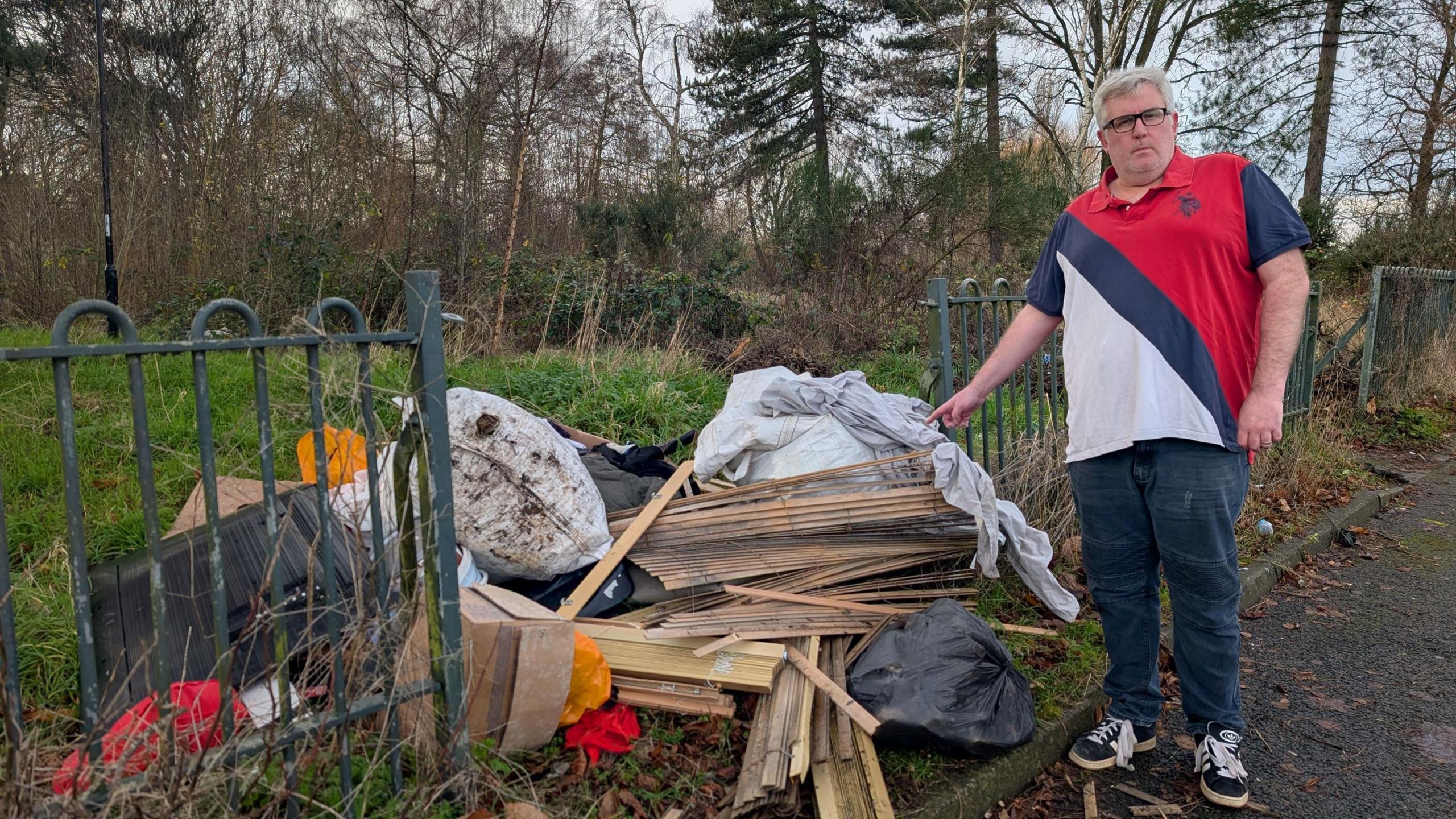 Frank Beechey, who is wearing a red, blue and white T-shirt, is standing on a path and pointing at a pile of waste that has been fly-tipped on a grass verge next to a metal fence. There are tall trees and greenery in the background.