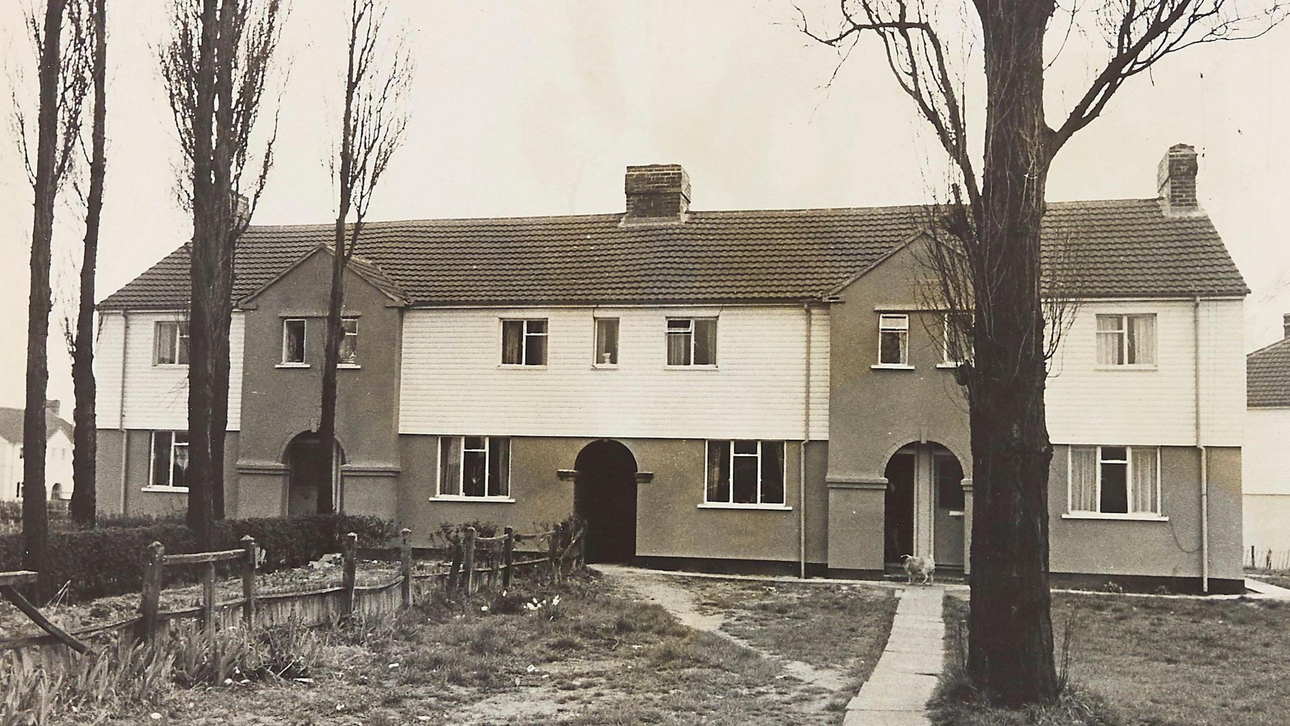 A black and white photo of terraced homes on two storeys. The homes are white at the top with brickwork on the bottom storey. They also have chimneys. In front of the homes are several trees and some grass areas with a wooden fence. 