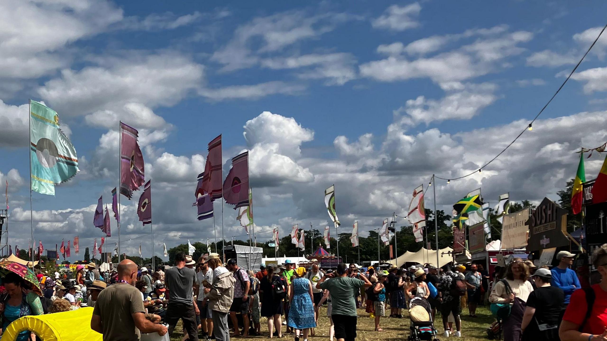 A festival crowd on a sunny day, with tall colourful flags on poles and tents lining the sides