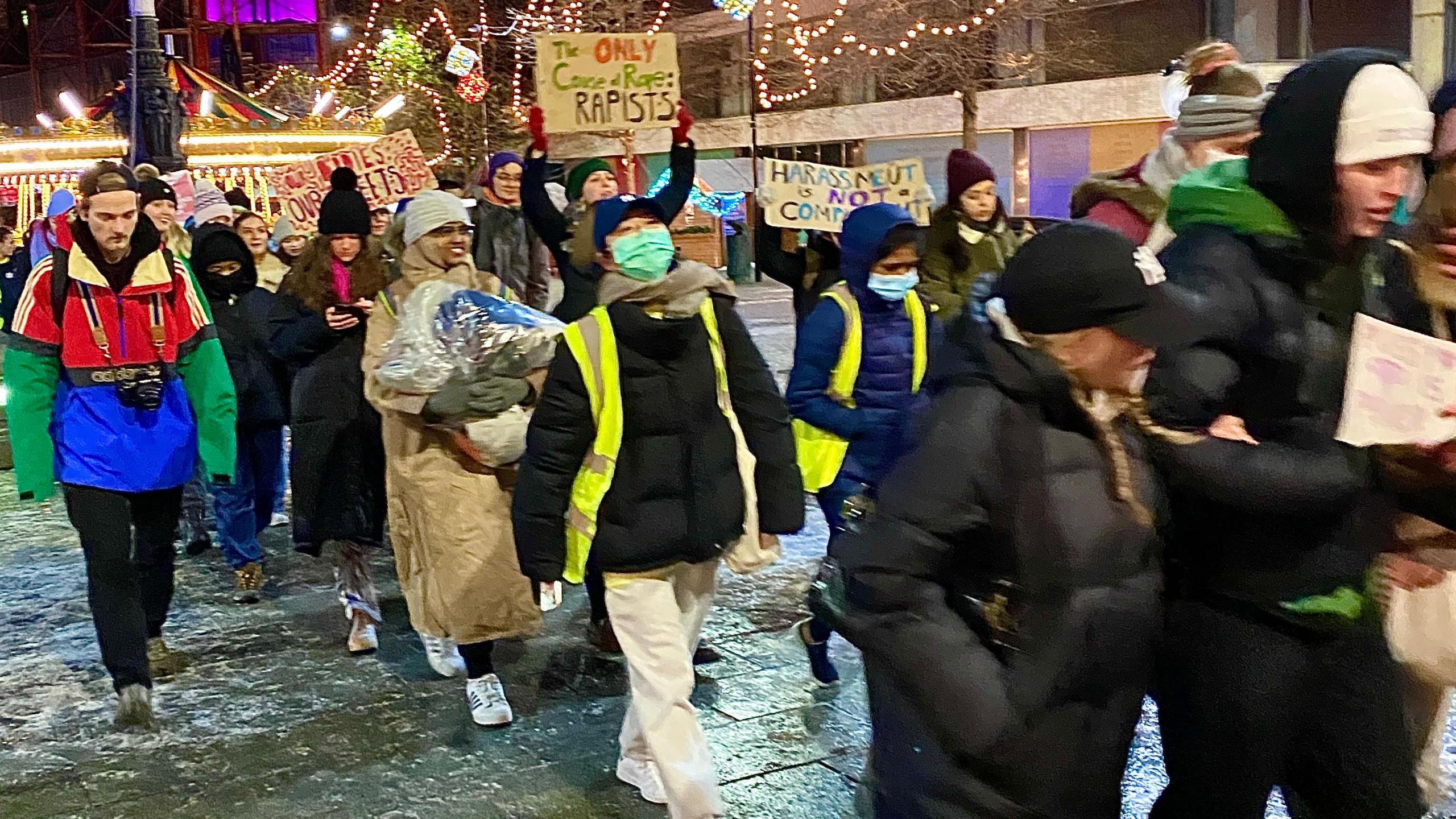 A crowd including men and women gathered in front of buildings lit by Christmas lights. Some are holding banners which read "The only cause of rape is rapists" and "Harassment is not a compliment".