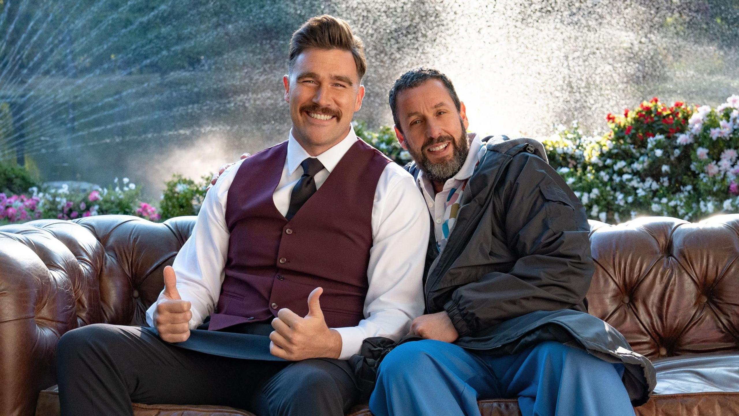 Travis Kelce with both thumbs up sitting on a leather sofa with Adam Sandler, both smiling, on the set of Happy Gilmore 2