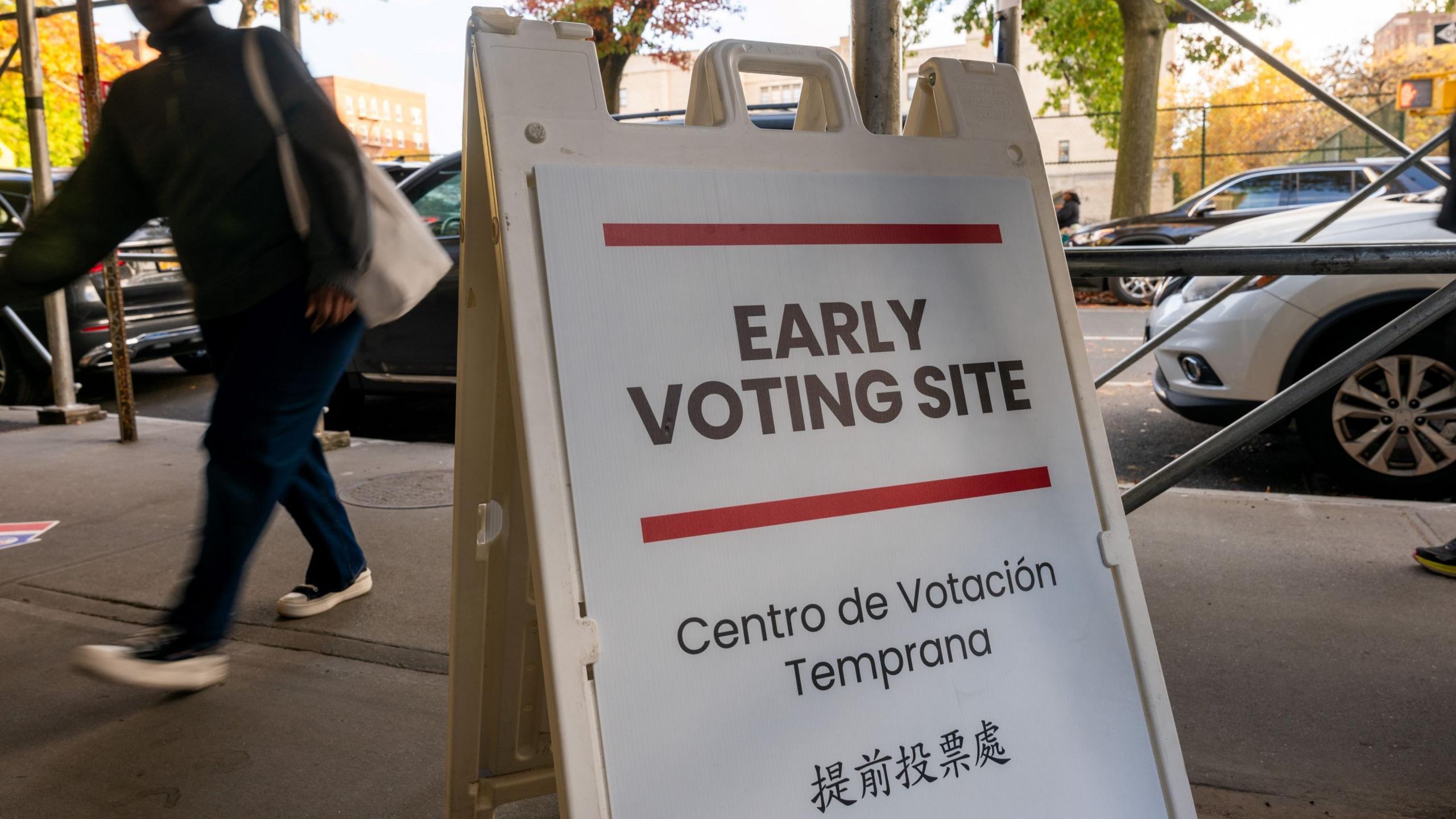 An unidentified person dressed in dark clothing walks past a sandwich-board style sign on a sidewalk. The sign reads "Early voting site" in several languages