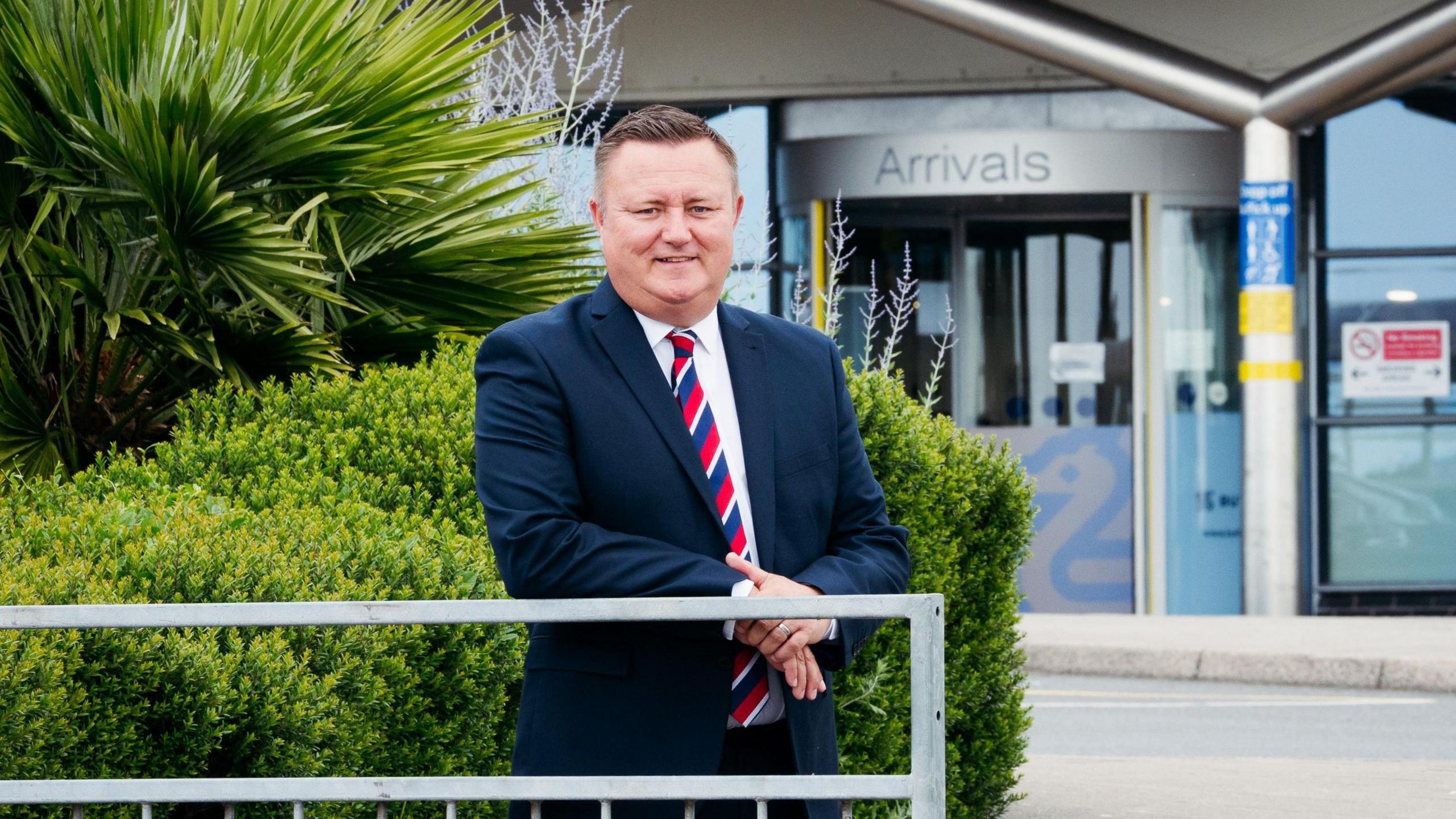 Richard Thomasson stands in a suit and tie smiling outside the airport, with the word Arrivals near him.