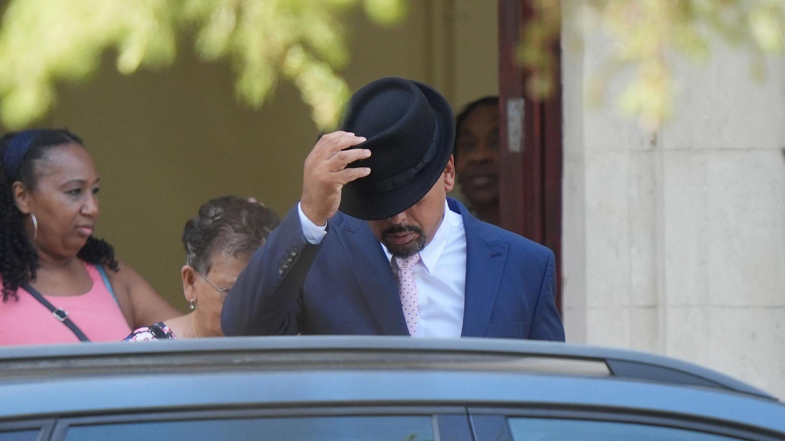 A man removing a black hat from his head. He is leaving a building while wearing a dark blue suit