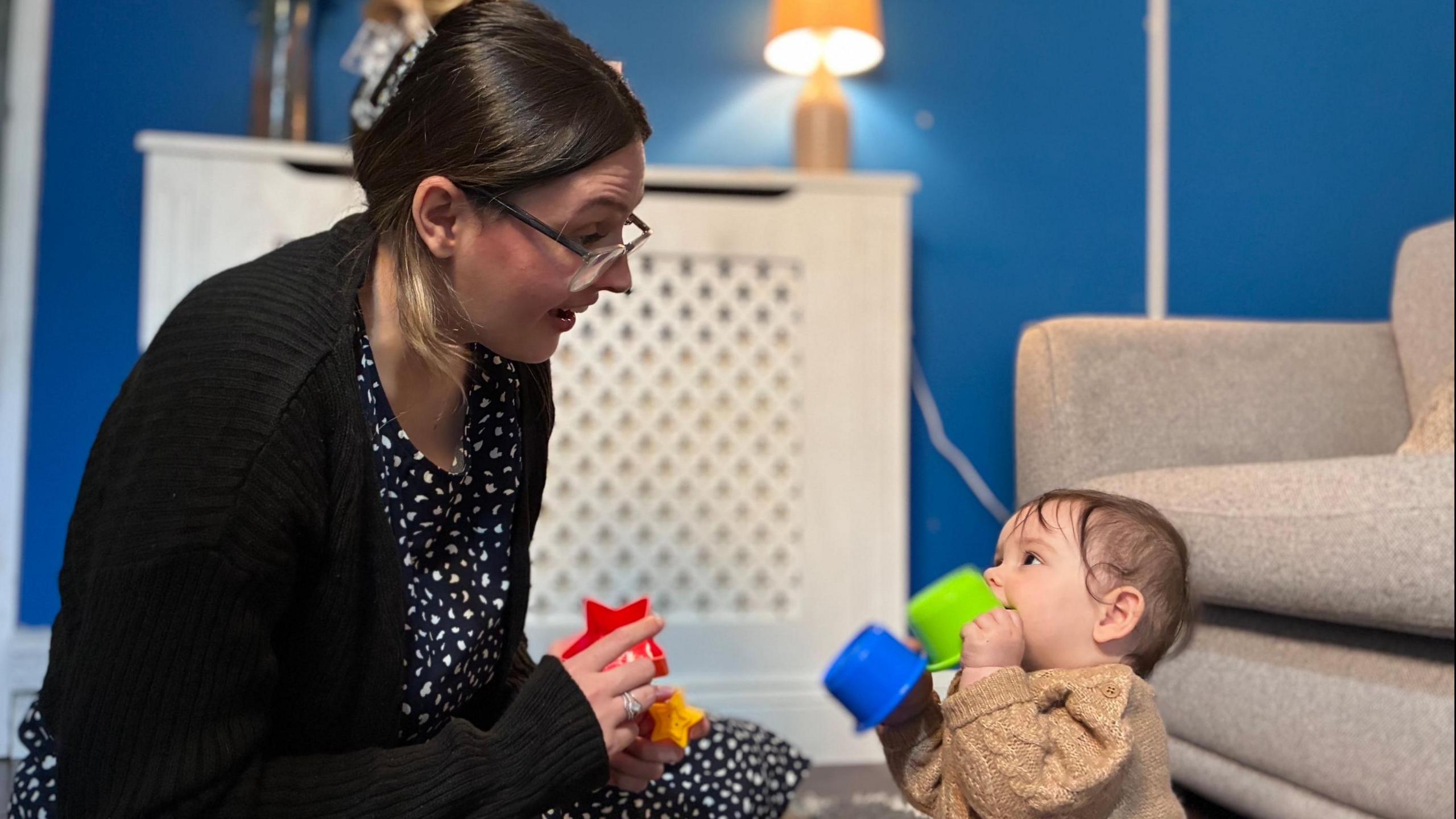 Mae Chloe a Daisy yn eistedd ar ryg yn eu stafell fyw. Mae Chloe, sy'n gwisgo ffroc glas a gwyn, sbectolau a cardigan du, gyda'i gwallt wedi ei clipio, yn tynnu gwynebau ar Daisy. Mae Daisy yn cnoi ar cwpan plastig gwyrdd ac yn edrych yn astud. ar ei mam. Mae'r ddau yn dal cwpanau plastig. Mae glas ar wailau'r stafell fyw, gyda gwresogydd gwyn, soffa a golau lamp yny. cefndir.